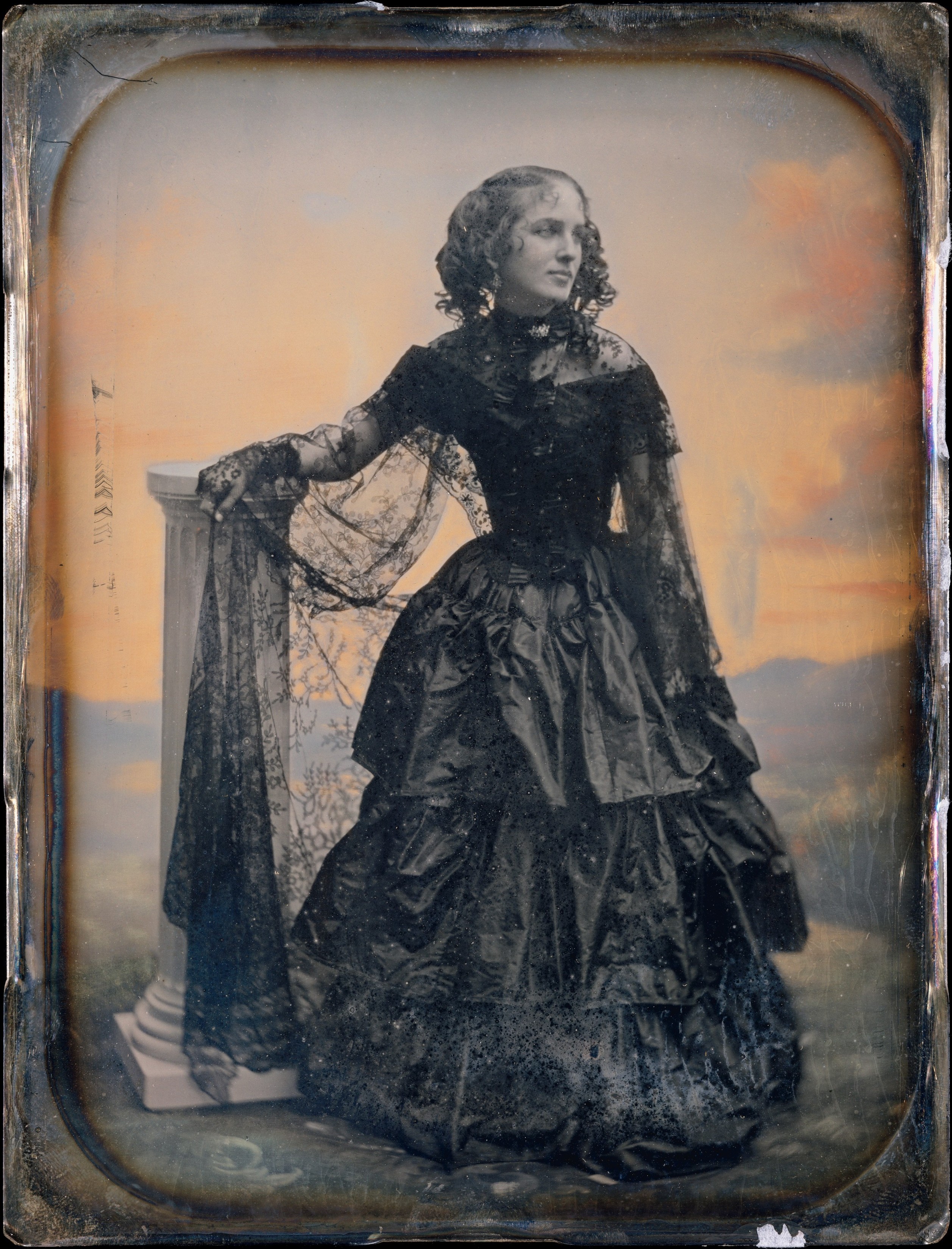 A vintage photograph of a white woman wearing a short sleeved dark taffeta dress, with a lace shawl and lace glove. Her right arm rests on a pedestal. The background has applied colors and resembles a landscape.