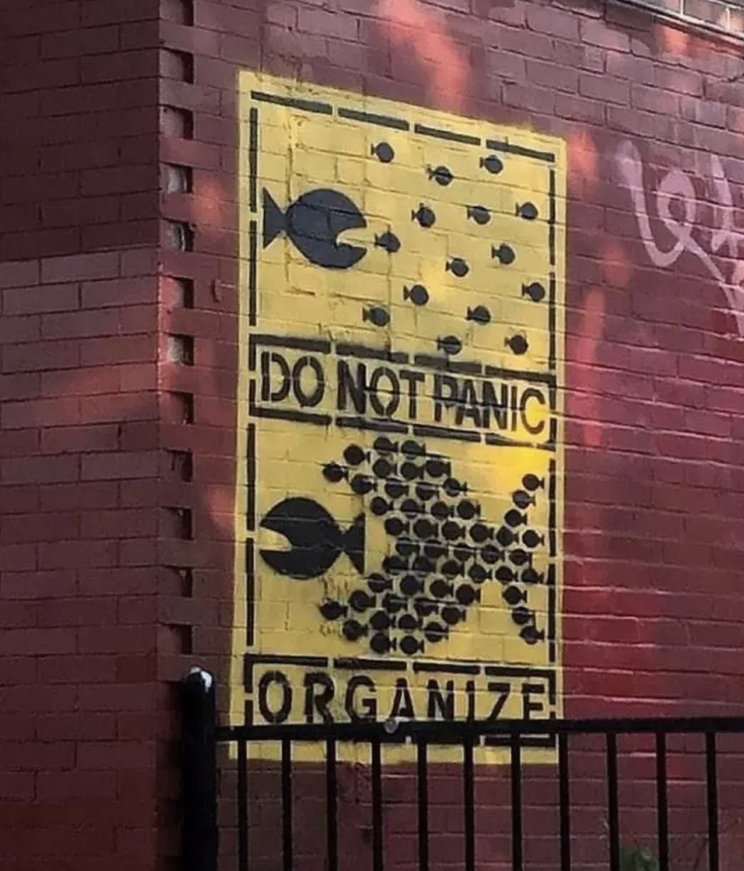 A stencil mural on a red brick wall in Providence, Rhode Island, USA. The artwork features a bold yellow background divided into two sections. The top shows a single large black fish surrounded by smaller fish scattered in disarray, with the text "DO NOT PANIC." The bottom section illustrates the smaller fish united to form a larger, powerful fish, swimming together to face the bigger fish, accompanied by the text "ORGANIZE." The piece delivers a powerful social message of unity and collective strength against challenges. A black fence runs along the bottom edge of the mural.