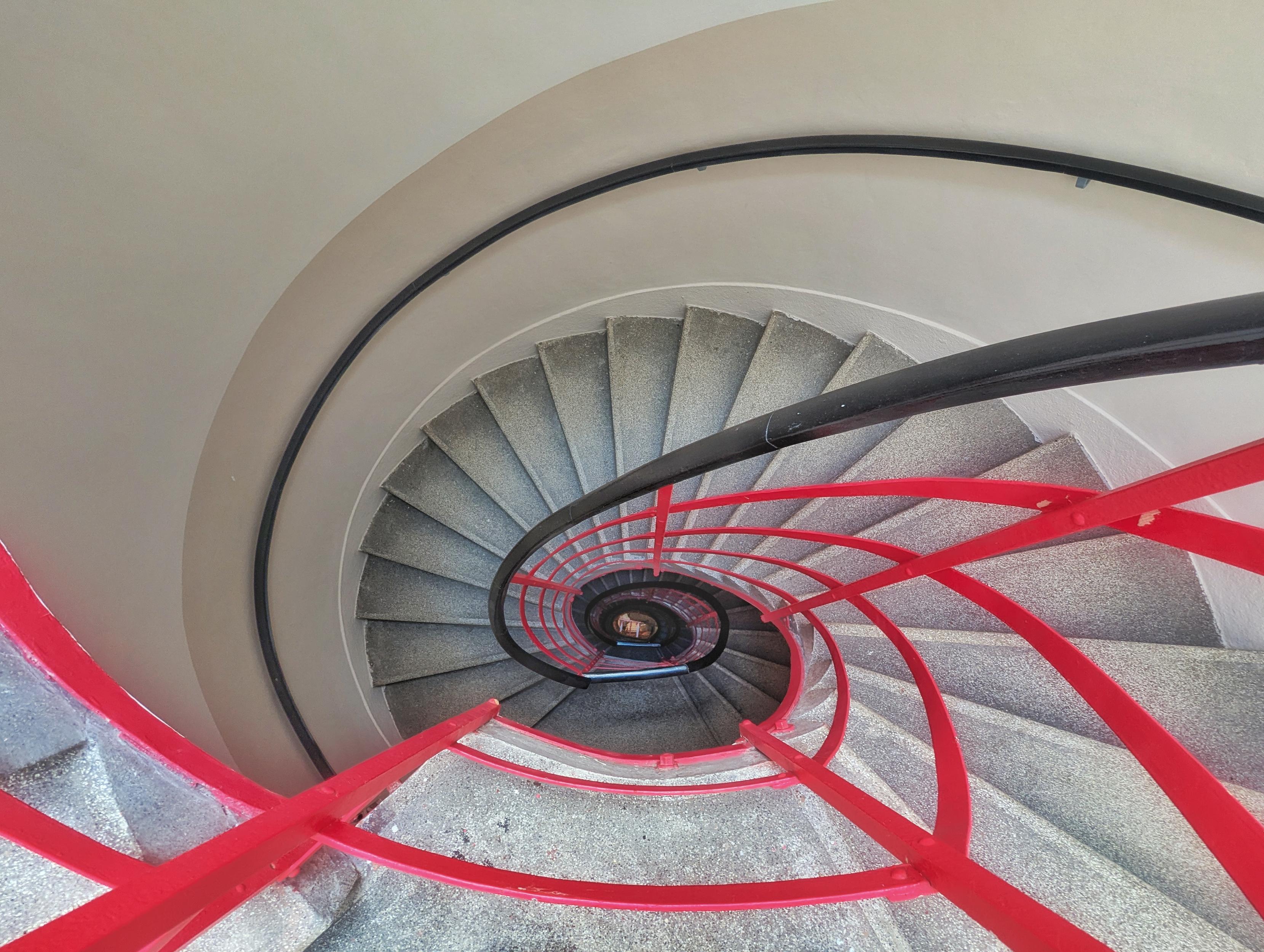 Top-down view of a modern spiral staircase with grey stone steps, a black handrail, and a bold red railing, winding through the centre of a multi-storey house.