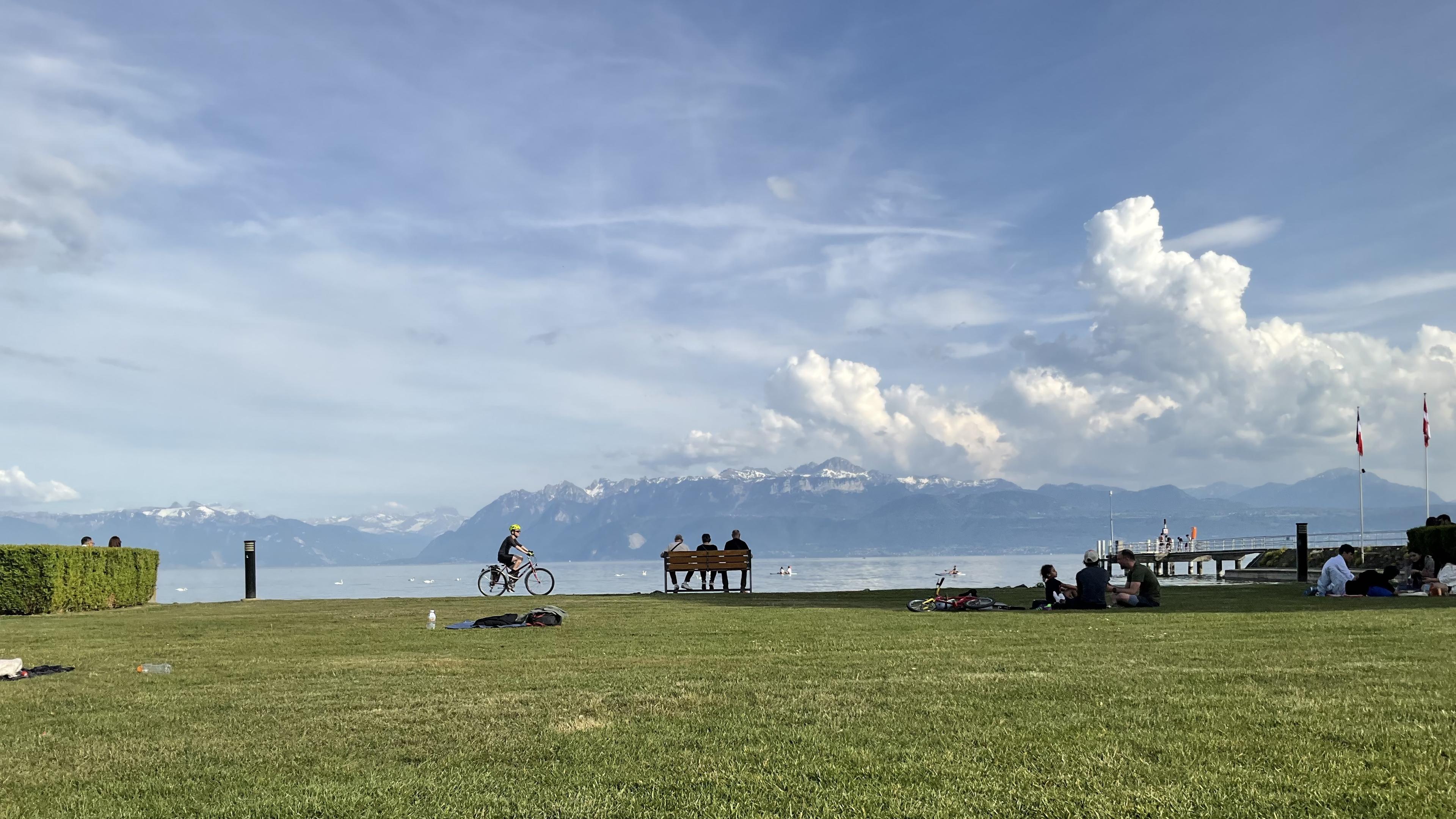 Vue du Léman. Cycliste, personnes sur un banc. Dans l’arrière-plan, les Alpes et des nuages.