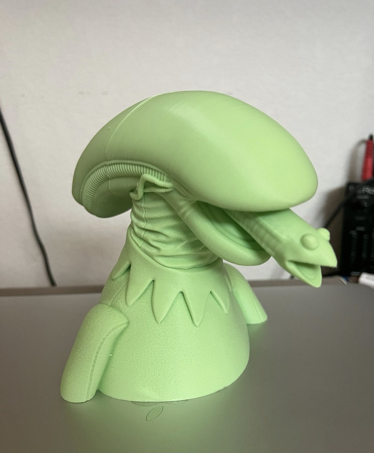 A 3d printed object that is the unholy hybrid of a xenomorph, and Kermit the Frog.  It is the green body of Kermit, with the long curved head of the xenomorph, and Kermit’s head popping out of the xenomorph’s mouth like the “little head” of the xenomorph. @NuclearOatmeal@beige.party says "In space, it isn't easy being green."