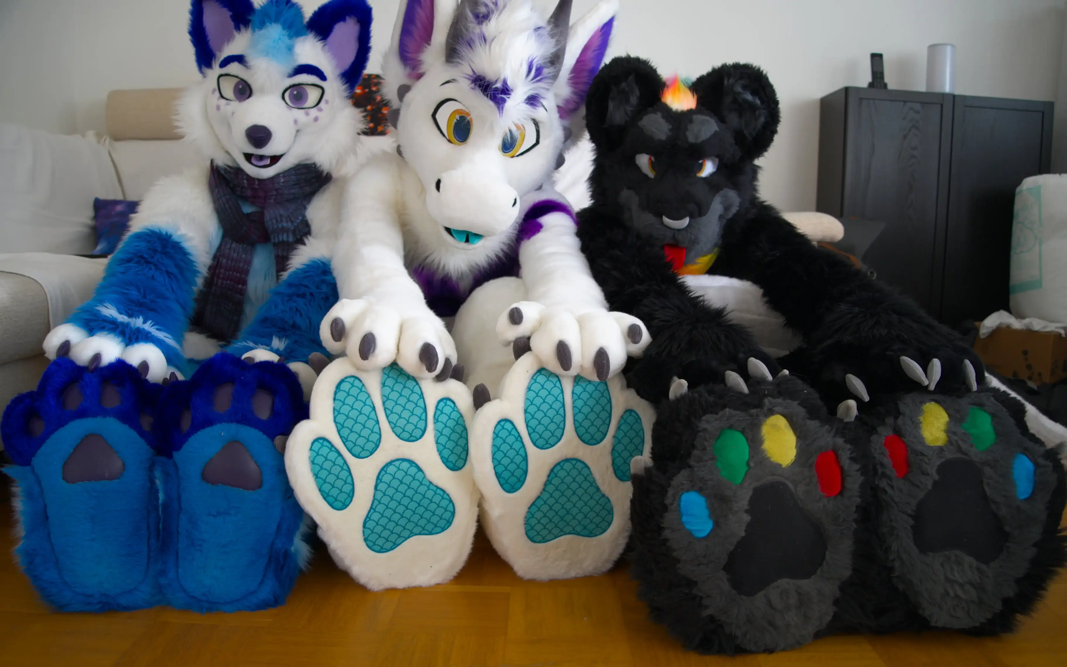 Photo of three fursuiters sitting on a wooden living room floor facing the camera and touching their foot paws: Polarie, an arctic fox with white and blue fur and dark blue paw pads; Karb, a dragon with white and purple fur and cyan paw pads with a scale pattern; and Kianga, a black jaguar with rainbow-colored paw pads.
