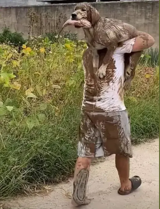 This photo shows a muddy dog being carried on a person's shoulder. The happy and very muddy dog appears to be holding a fish in its mouth, seemingly after a day spent playing in the mud... or?
