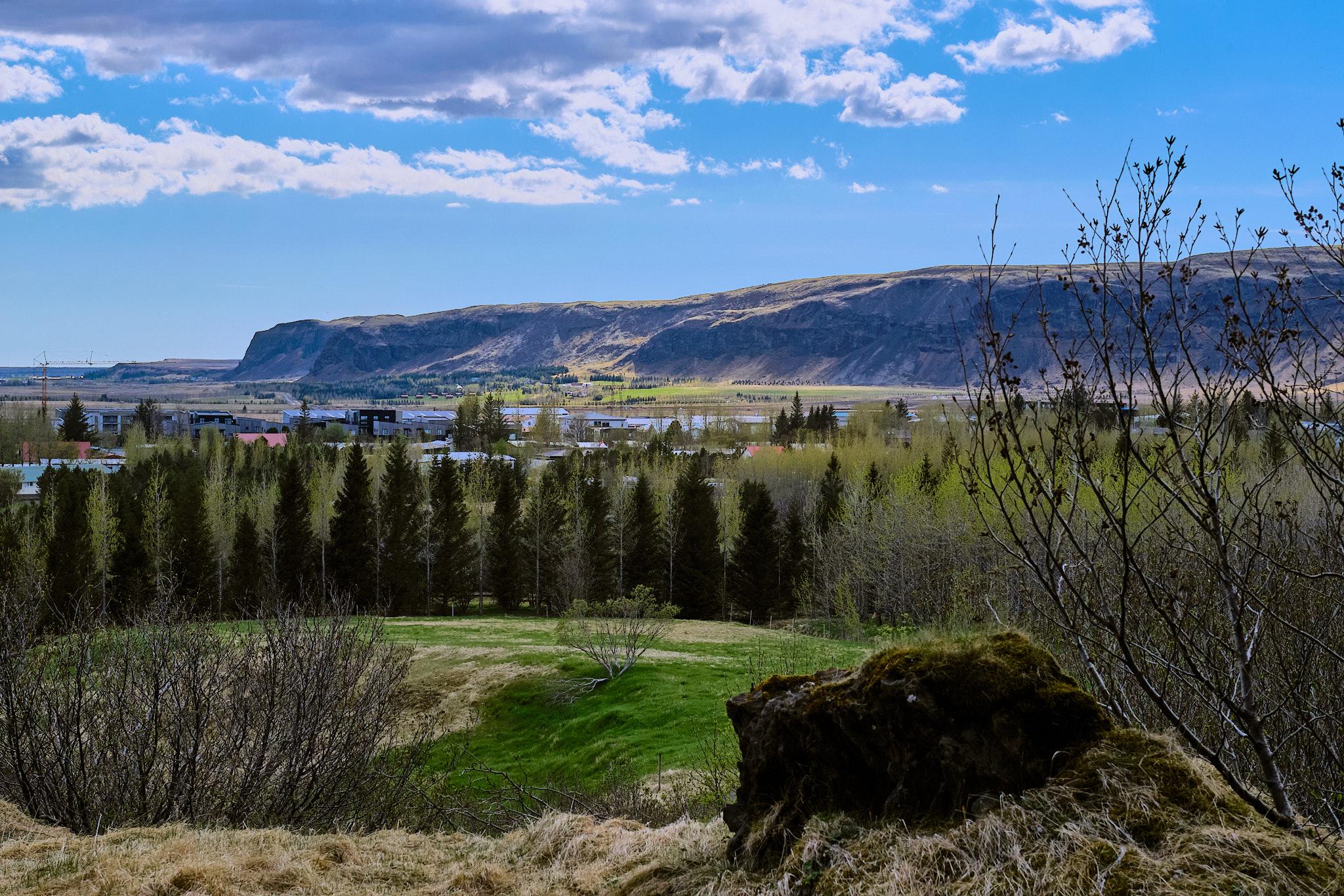 A view of Hveragerði town from the roots of Hverfjall mountain. You can see trees, mountains, and a blue sky