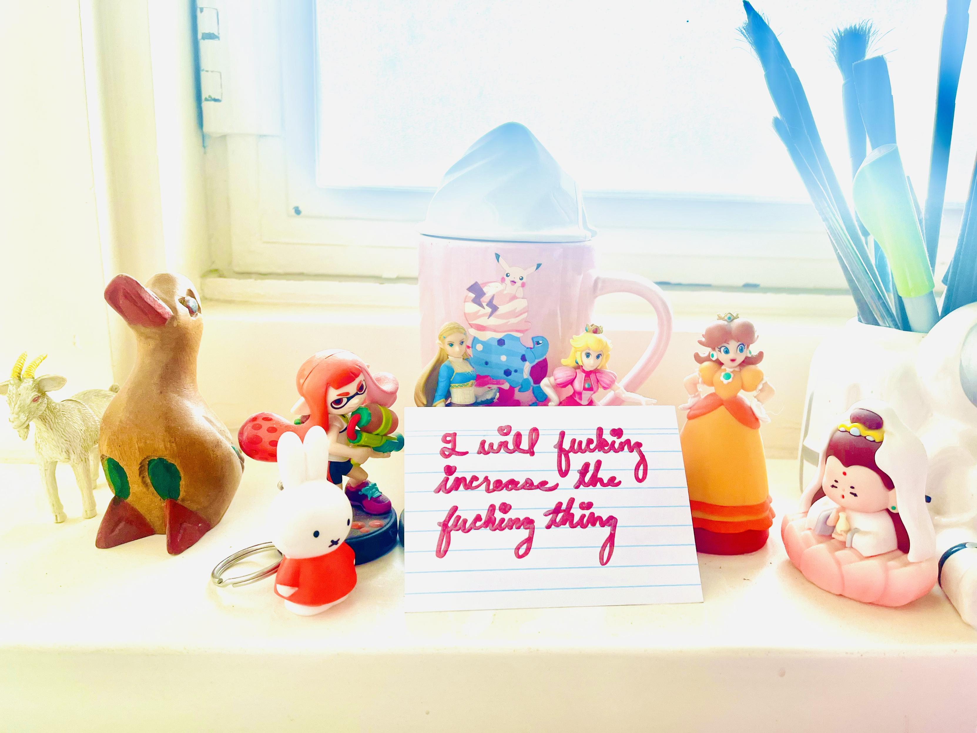 Handwritten pink glitter writing with heart-dotted i’s: “I will fucking increase the fucking thing.” The card is held up by several cute figurines: Nijntje (Miffy), Inkling Girl, Zelda, Peach, Daisy, and uhhh the goddess Guanyin