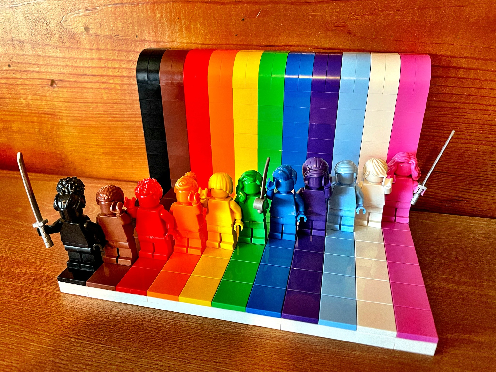 A rainbow-colored LEGO display set on a wooden surface. The display features a series of vertical colored blocks arranged in a spectrum from black to pink, resembling a Pride flag. In front of each colored section stands a matching monochrome LEGO minifigure. The black and pink figures at the ends are holding large swords, while the green figure in the middle holds a cutlass.