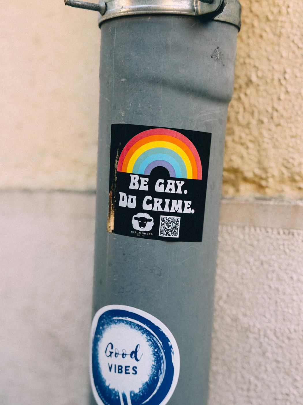A gray metal pole with two stickers, one featuring a rainbow and the text “Be Gay. Do Crime.”, and another circular blue and white sticker reading “Good Vibes.”