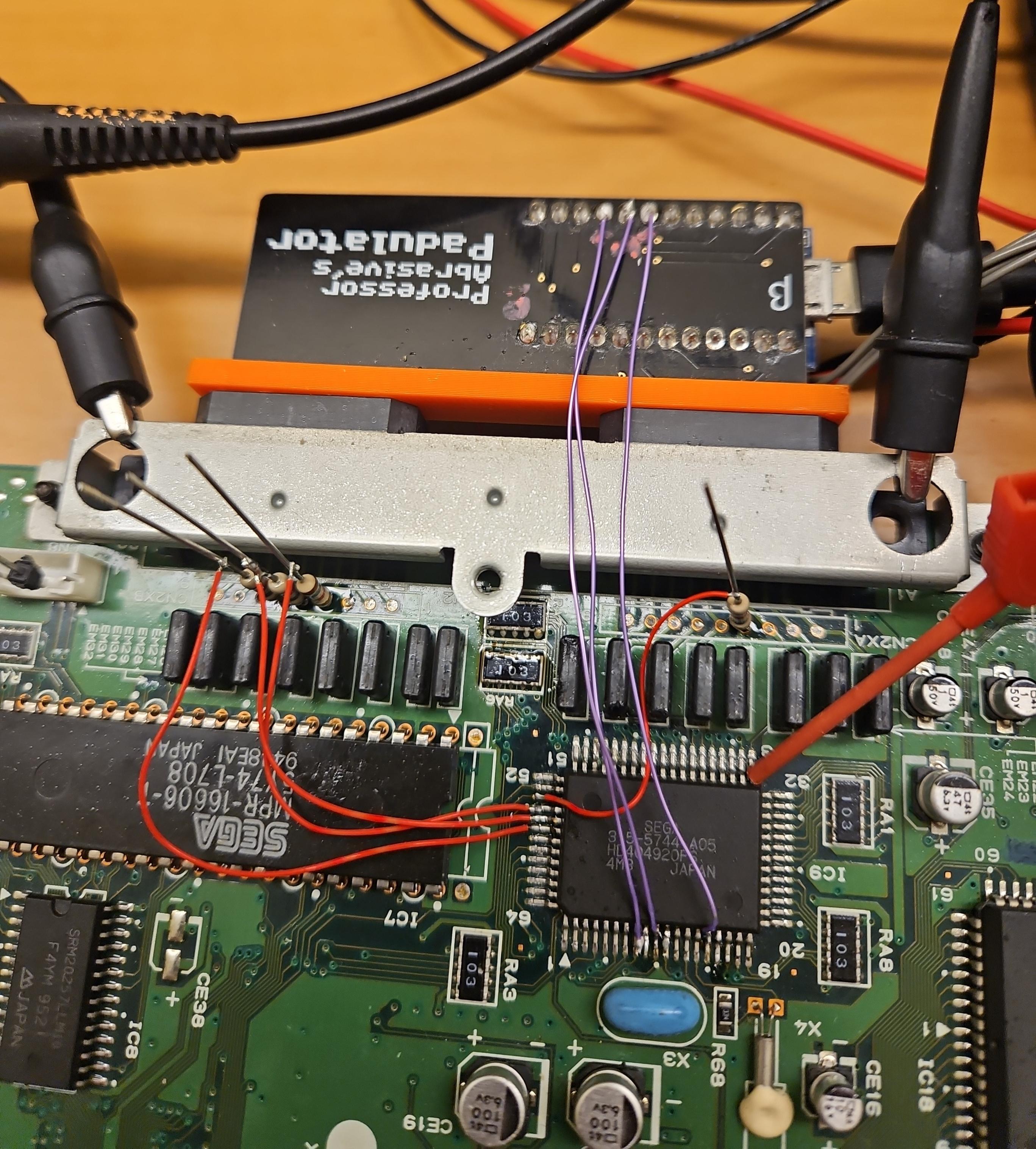 The mainboard of a Sega Saturn. Diverse wires have been attached to one particular chip on the board. Some of them run to a doover which is plugged into both controller ports - labelled Professor Abrasive's Padulator - from which a USB cable disappears out of frame. A couple of the chip's legs have been lifted from the board.