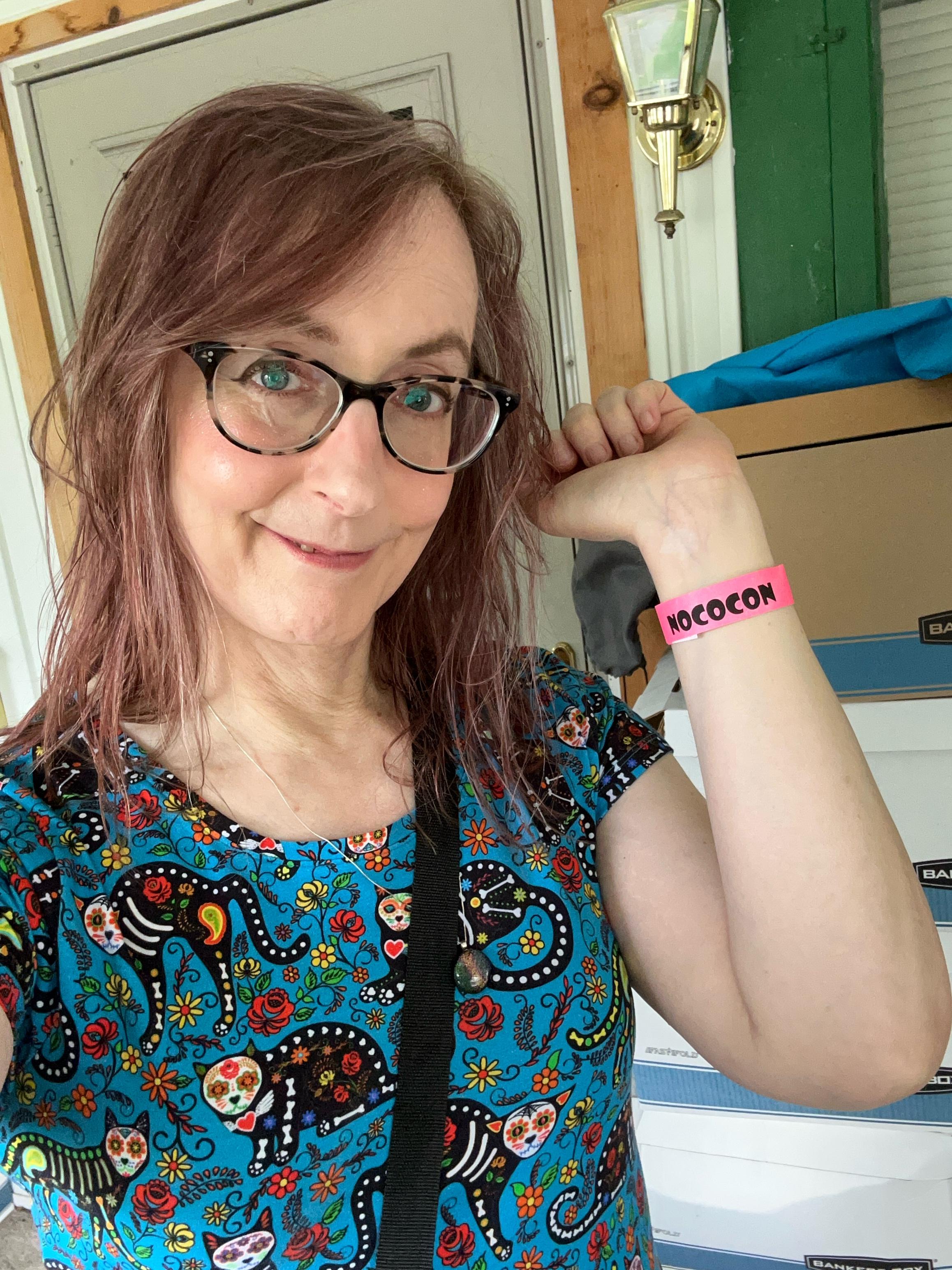 An adult white woman with long brown hair and gray eyes, wearing tortoiseshell cat eye glasses and a dress with sugar skull cats with skeletal bodies frolicking amidst roses and marigolds against a turquoise background. She is smiling at the camera and holding up her left wrist to show a pink band that says "Nococon."