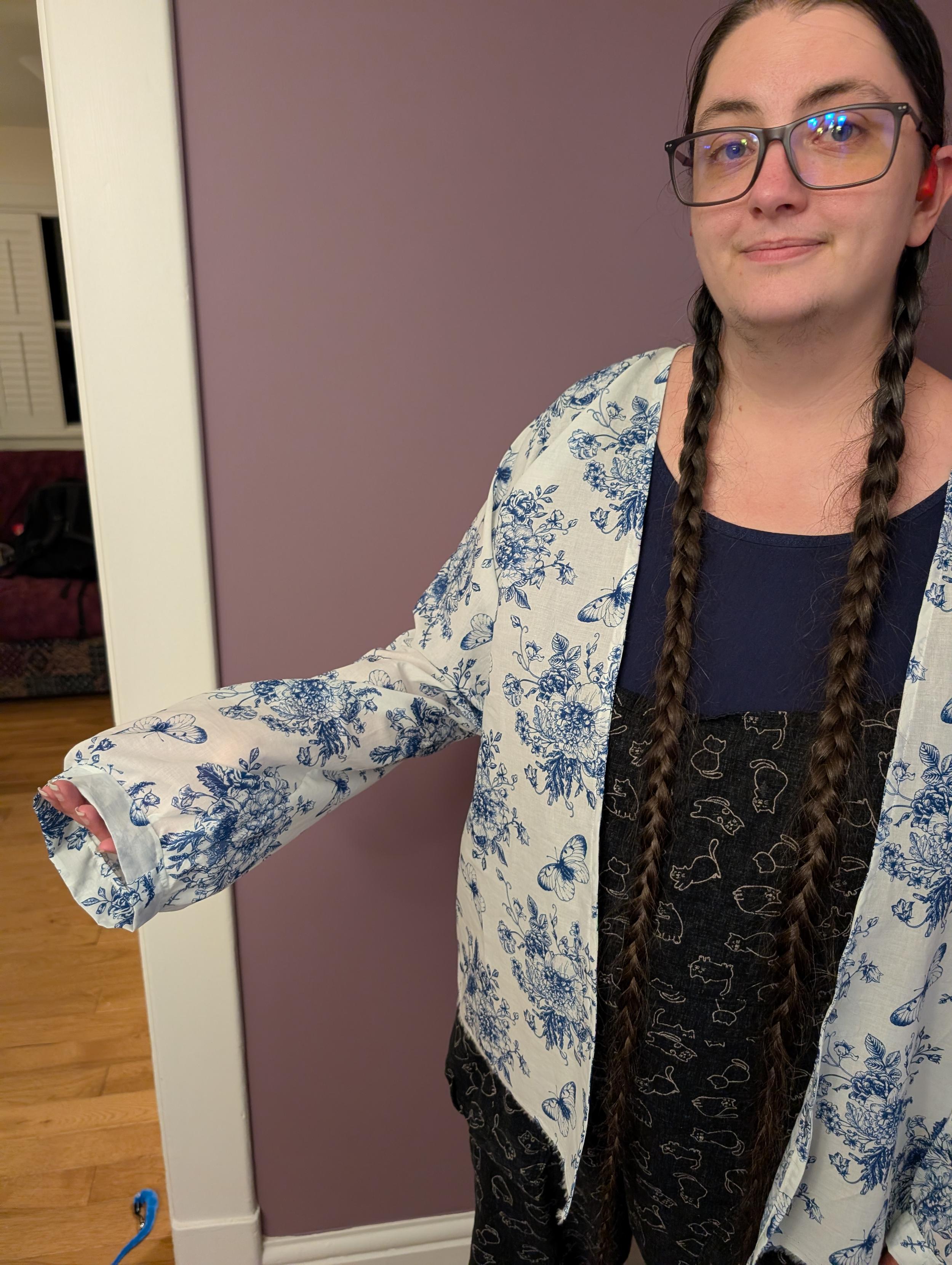 A photo of myself wearing a white muslin overshirt with a blue pattern of butterflies and flowers over overalls with a cat print and a blue tee shirt.