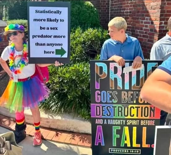 A person wearing a rainbow tutu, lei, and socks stands next to a religious demonstrator. The rainbow-clad person holds a sign with a green arrow pointing to their right that reads: "Statistically more likely to be a sex predator more than anyone here". The arrow points directly at a young man holding a large black sign with rainbow lettering that quotes Proverbs 16:18: "PRIDE GOES BEFORE DESTRUCTION AND A HAUGHTY SPIRIT BEFORE A FALL."