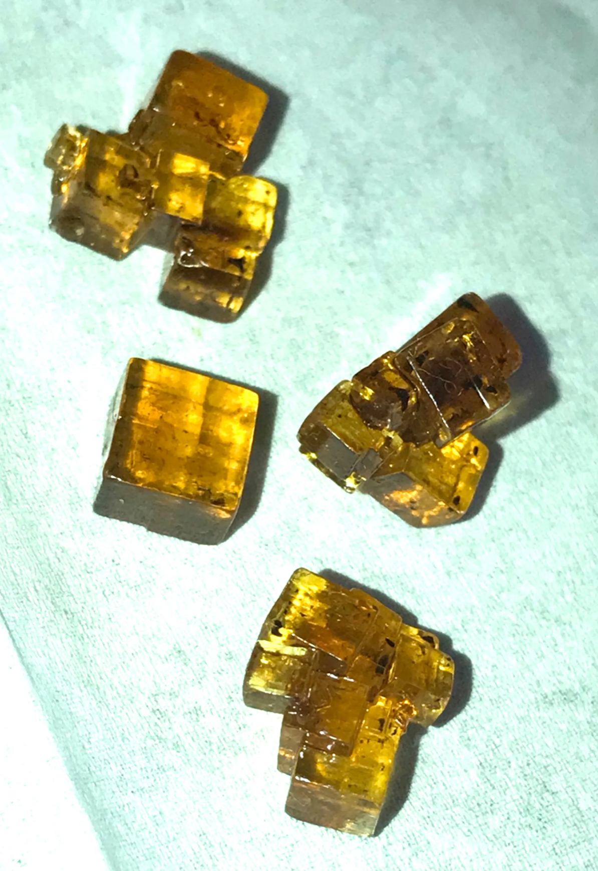 A photo of four large salt crystals, golden coloured and like little cubes of transparent gold. Three of them are groups of cubes joined together, and one is a single slightly rectangular cube by itself, all with wonderfully ninety degree angles on them.
