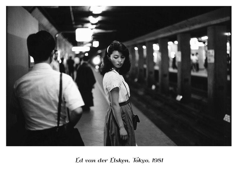 The image is a black and white photograph depicting a scene from Tokyo in 1981, as noted by the text at the bottom. It captures a moment on what appears to be an underground subway platform or similar transportation hub. In the foreground stands a young woman with shoulder-length hair wearing a light-colored blouse tucked into a high-waisted skirt and carrying a dark bag over her right shoulder. She is looking away from the camera, possibly down at something in her hand, conveying a sense of contemplation or distraction. Behind her to the left is another individual, presumably male, viewed from behind as he moves along the platform. He wears a white shirt with rolled-up sleeves and has dark hair. The setting features structural elements such as pillars supporting overhead beams, indicative of an enclosed transit area designed for pedestrian traffic above tracks below ground level. Various figures are visible in the background but remain indistinct due to motion blur or distance. The overall composition emphasizes mood over clarity; it's a moment frozen in time that invites viewers to ponder what might have captured this woman's attention and evoke curiosity about her story within an urban context from nearly four decades ago.