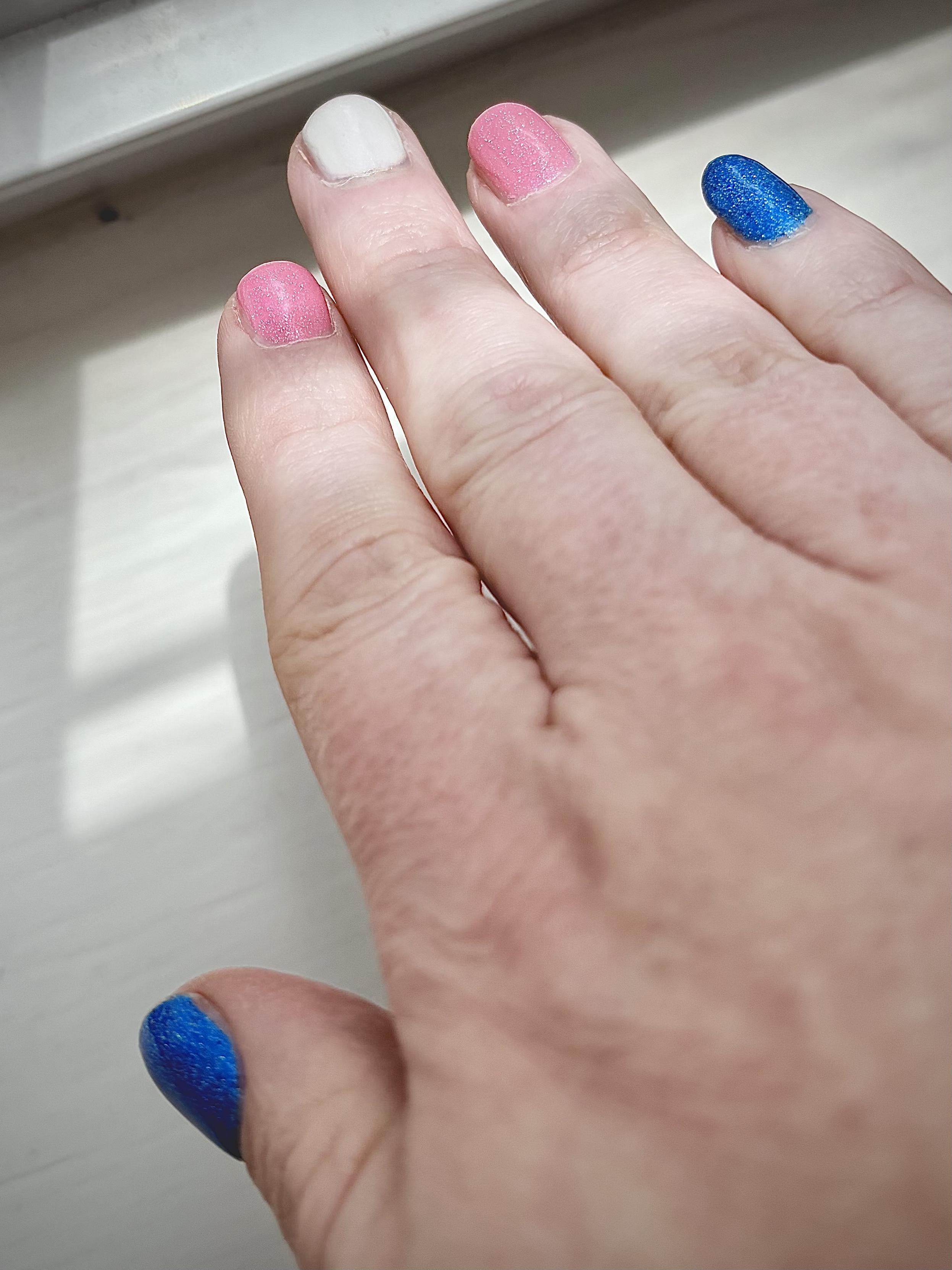 A hand with fingernails painted blue, pink, and white in the style of the trans pride flag.