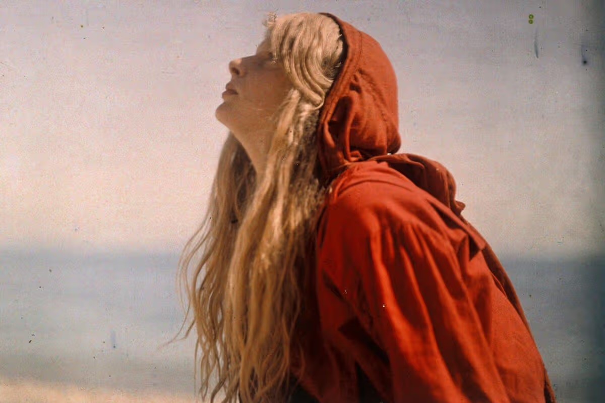 A head and shoulder length profile portrait photograph of a young woman  wearing a red cloak with a hood. Her long blond hair spills out from the hood. In the background, we see a beach.