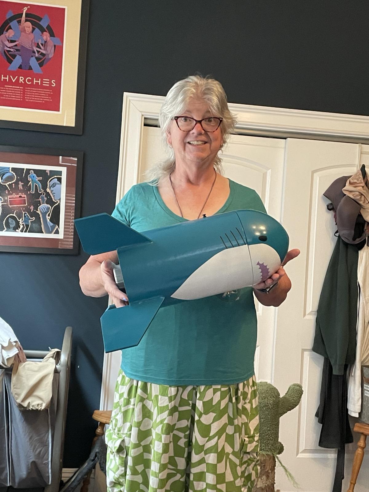 A smiling white woman with grey hair and glasses holding up a stubby, four fin model rocket decorated like a Blåhaj
