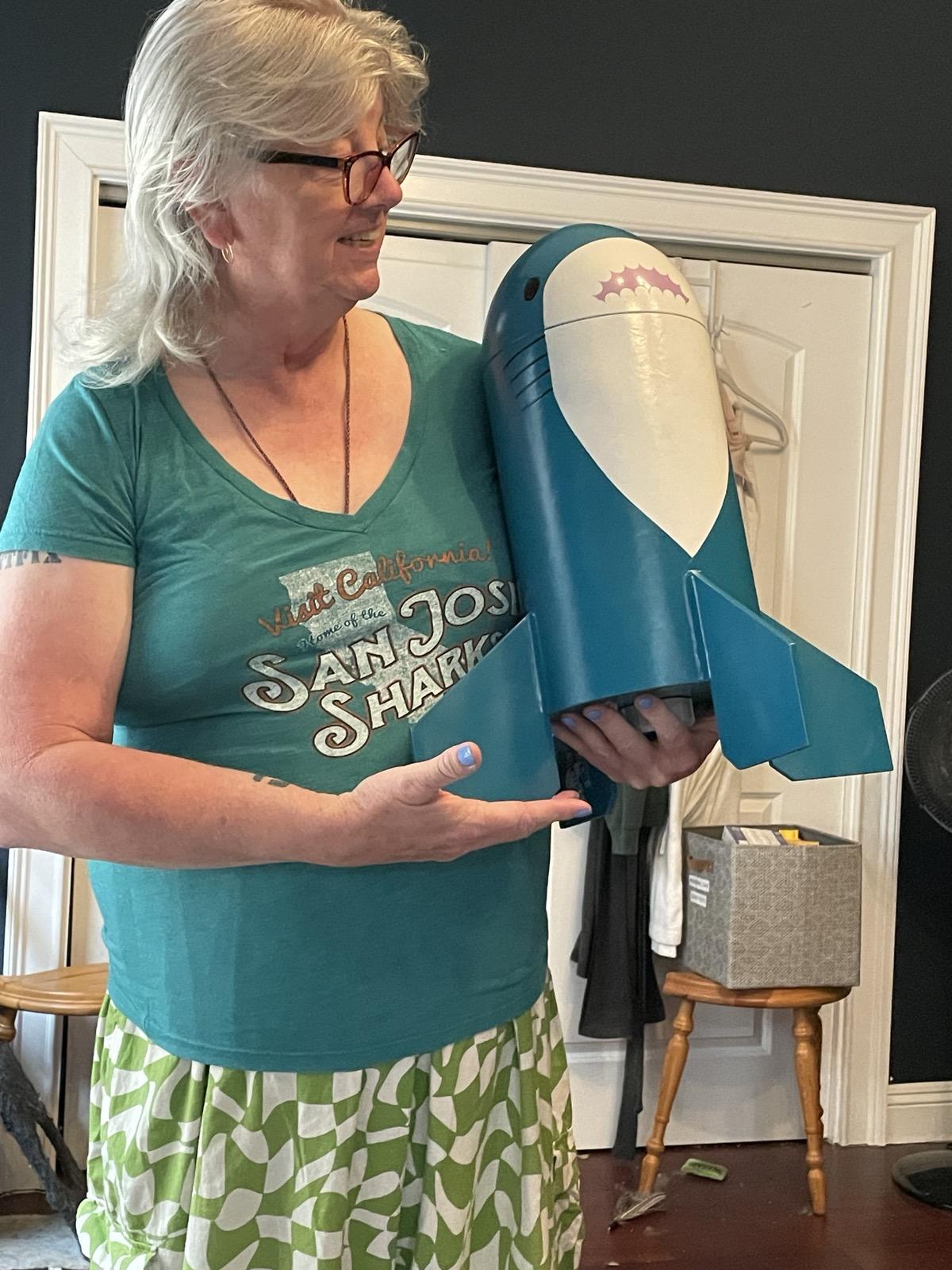 A smiling white woman with glasses holding a model rocket, decorated like a Blåhaj, like a small child