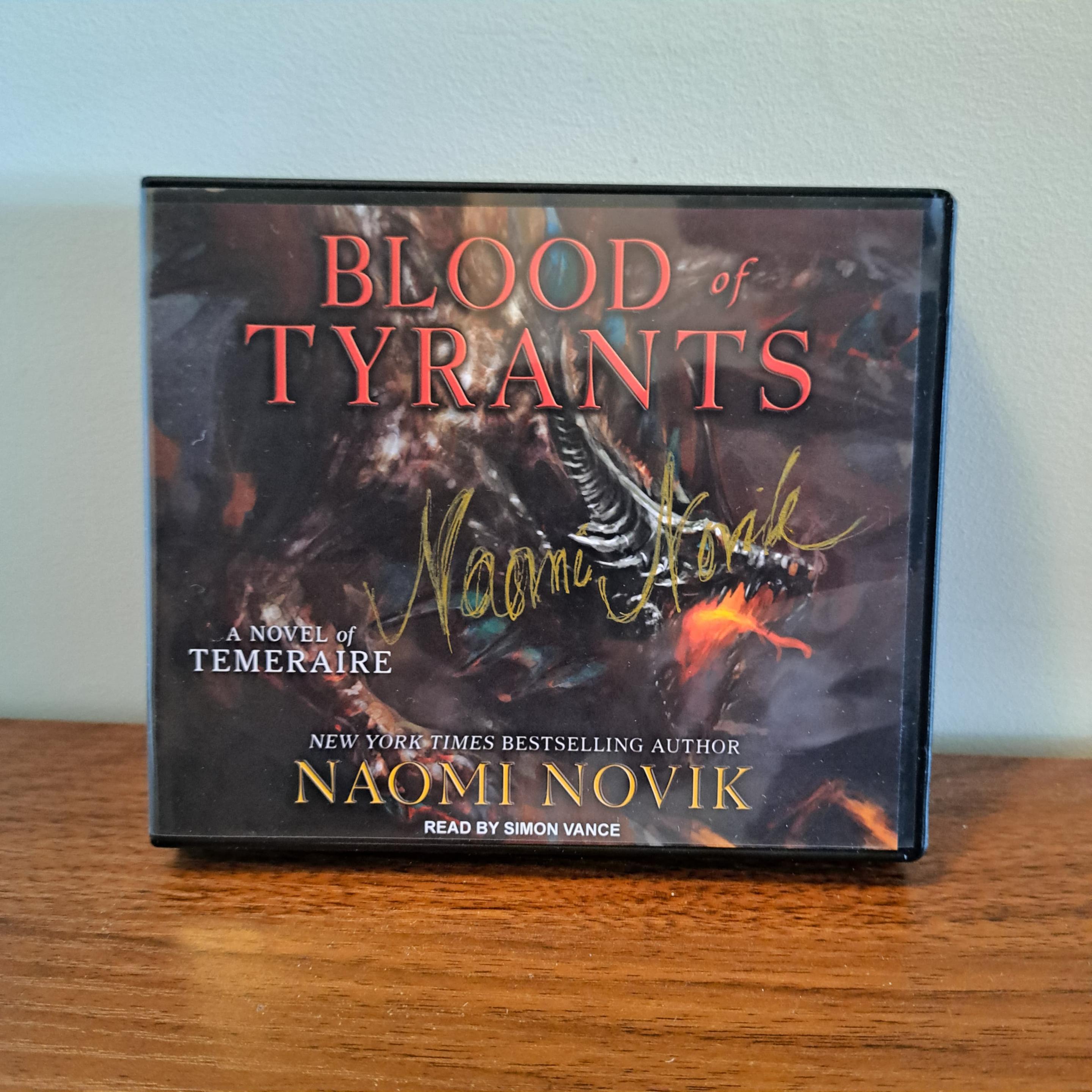 A boxed CD of BLOOD OF TYRANTS sitting on a wooden surface against a plain white background. The cover depicts the head, upper back and leg of a roaring reddish brown dragon with black spikes, emitting a bright reddish orange flame from wide open jaws. Title is orange-red in all caps across the top; author’s name is in dark yellow across the bottom, with a small tagline in white on the far left bottom, reading “A NOVEL OF TEMERAIRE.”  Naomi Novik’s signature in yellow marker is signed across the CD plastic casing.