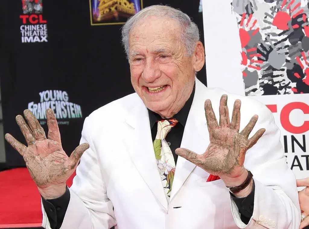 Mel Brooks holding up his hands after putting them in the wet cement and smiling for the camera. You can see a fake extra finger on his left hand
