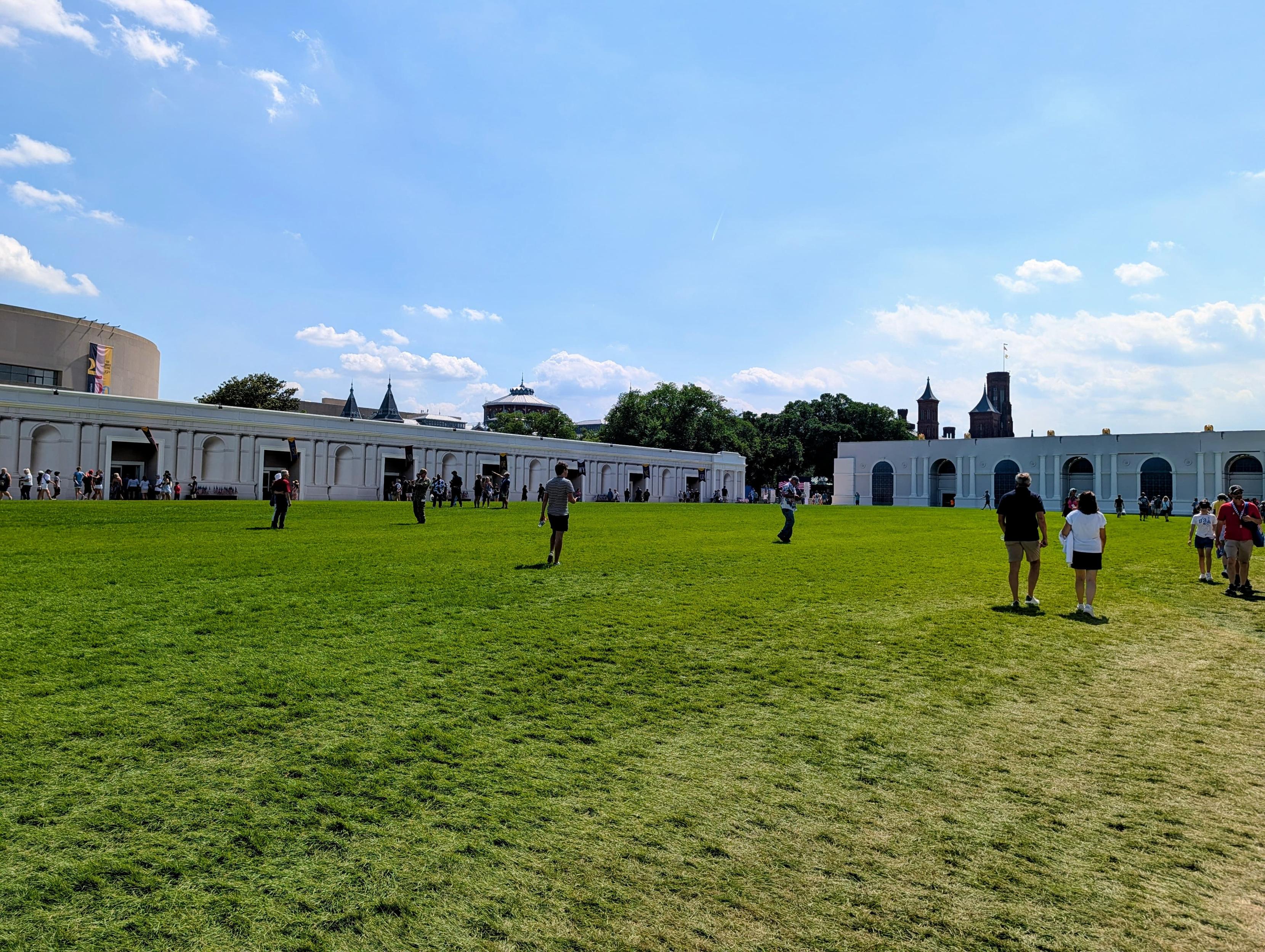 A large grassy expanse of the National Mall with a few people milling around. There are plywood sheds with poorly applied classical elements in the distance at the edge of the grass.