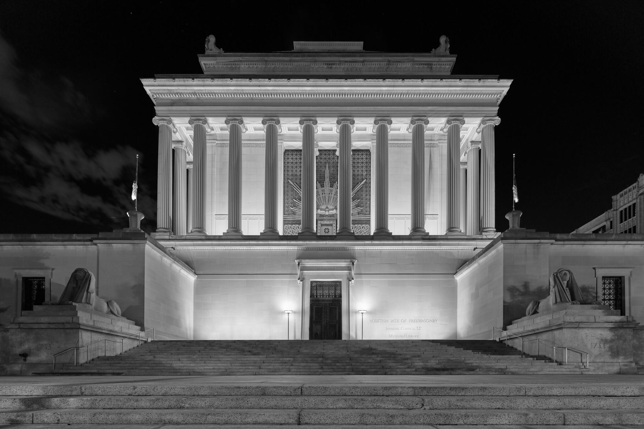 An imposing Masonic temple at night, with Ionic columns, ornate features, flanked by human lions statuary.