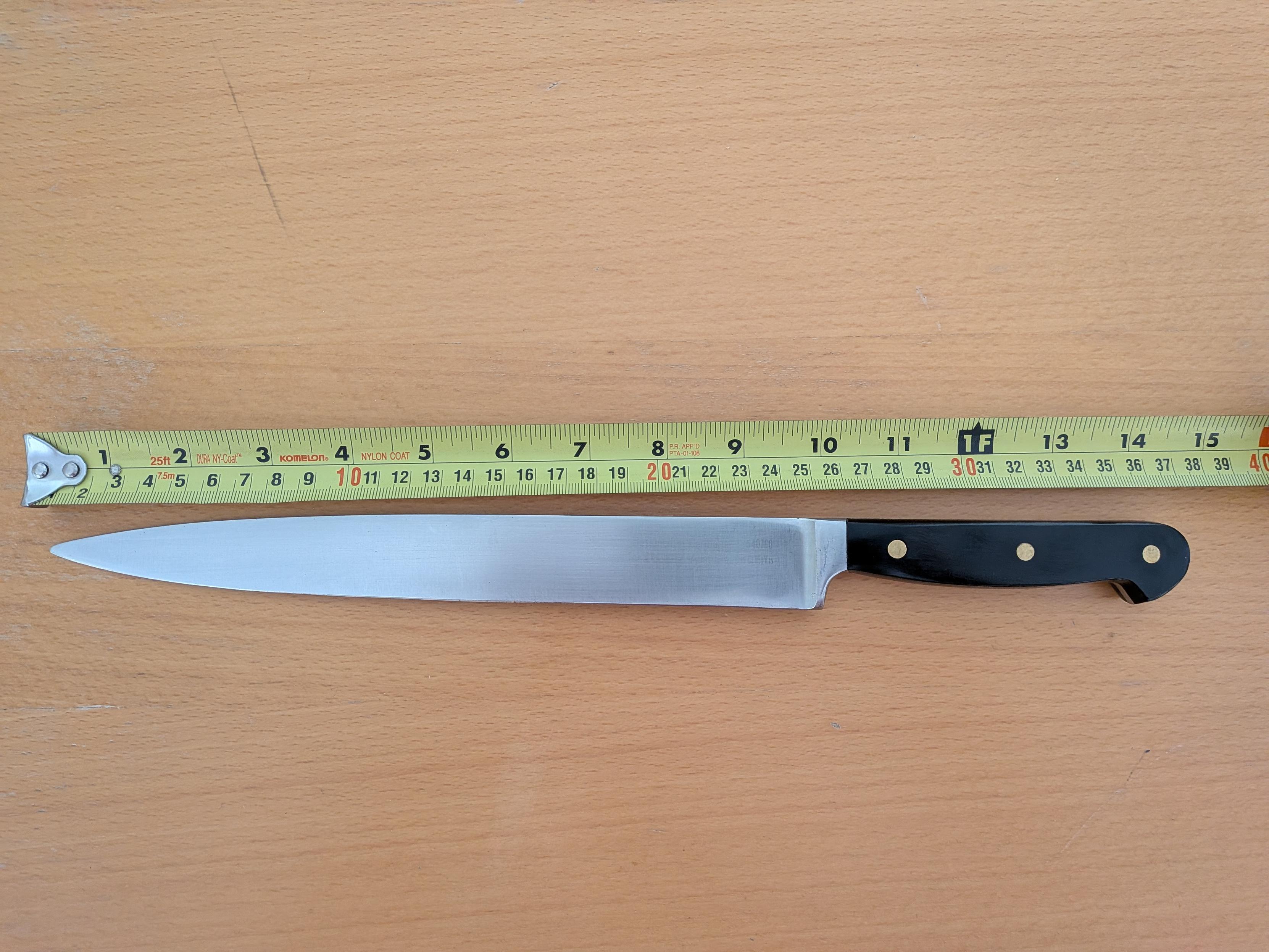 A kitchen knife on a table with a tape measure extended from the tip. The tape measure reads approx 10 inches/26cm where the blade connects to the handle.