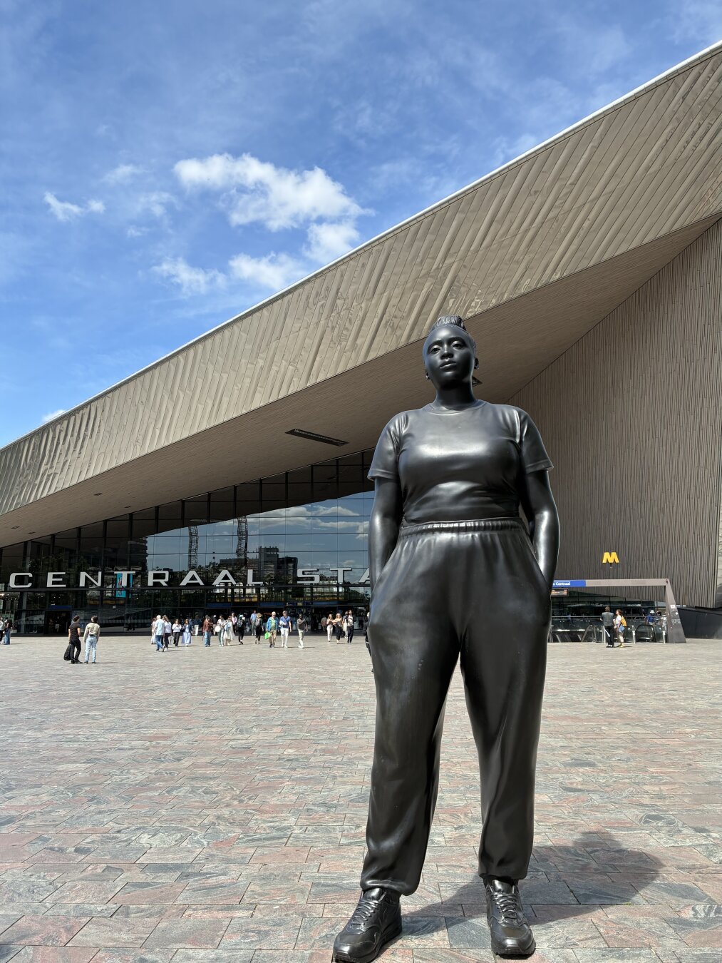 A large black statue of a woman stands in front of a modern building labeled "CENTRAAL STATION." The statue is dressed in casual attire and has hands in her pockets, while some people is visible in the far background, under a blue sky.