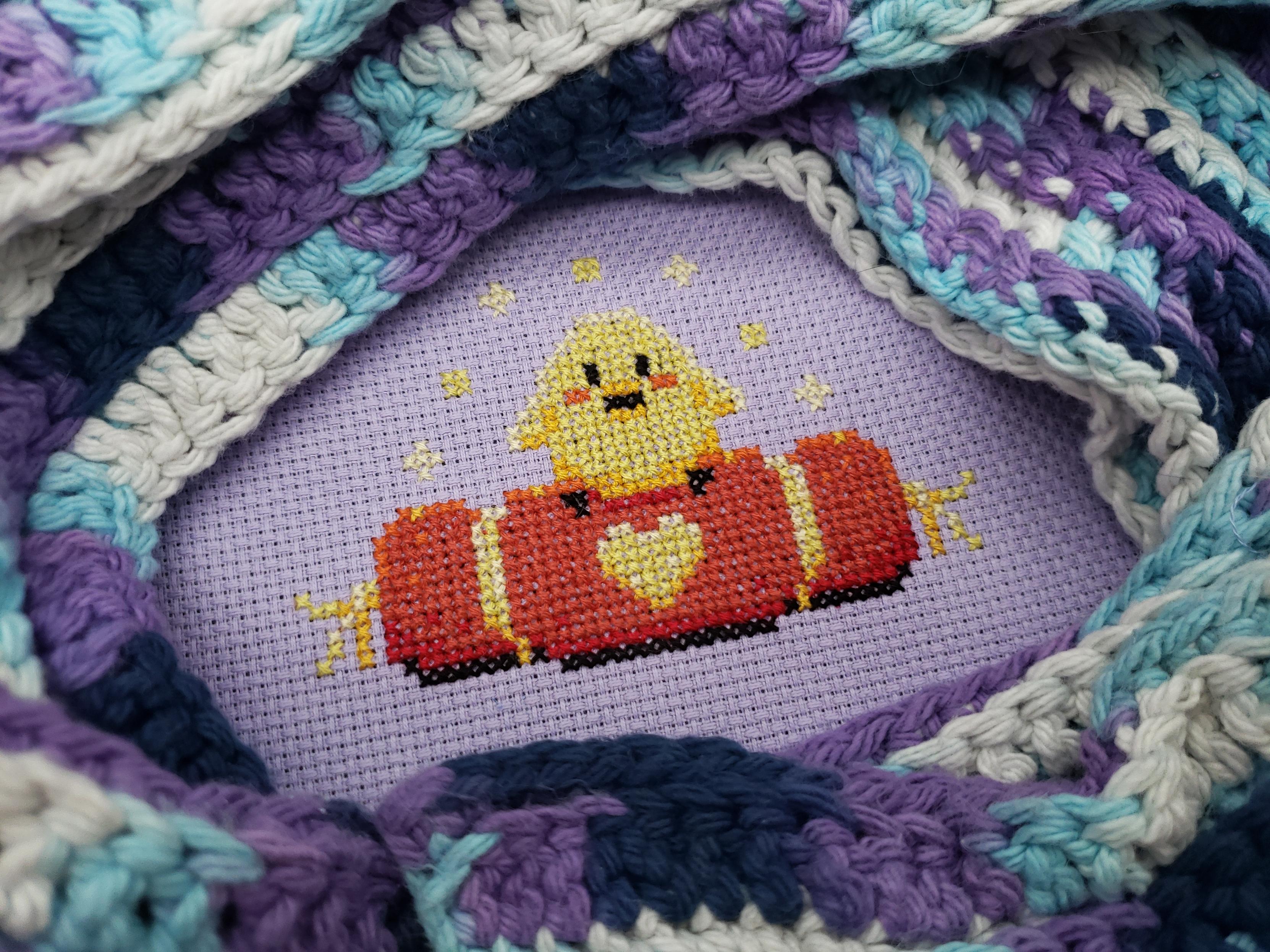 A cross stitch on light purple aida of a small chick sitting on an ornate red cushion. The cross stitch is framed by crocheted blue, purple, and white scarf.