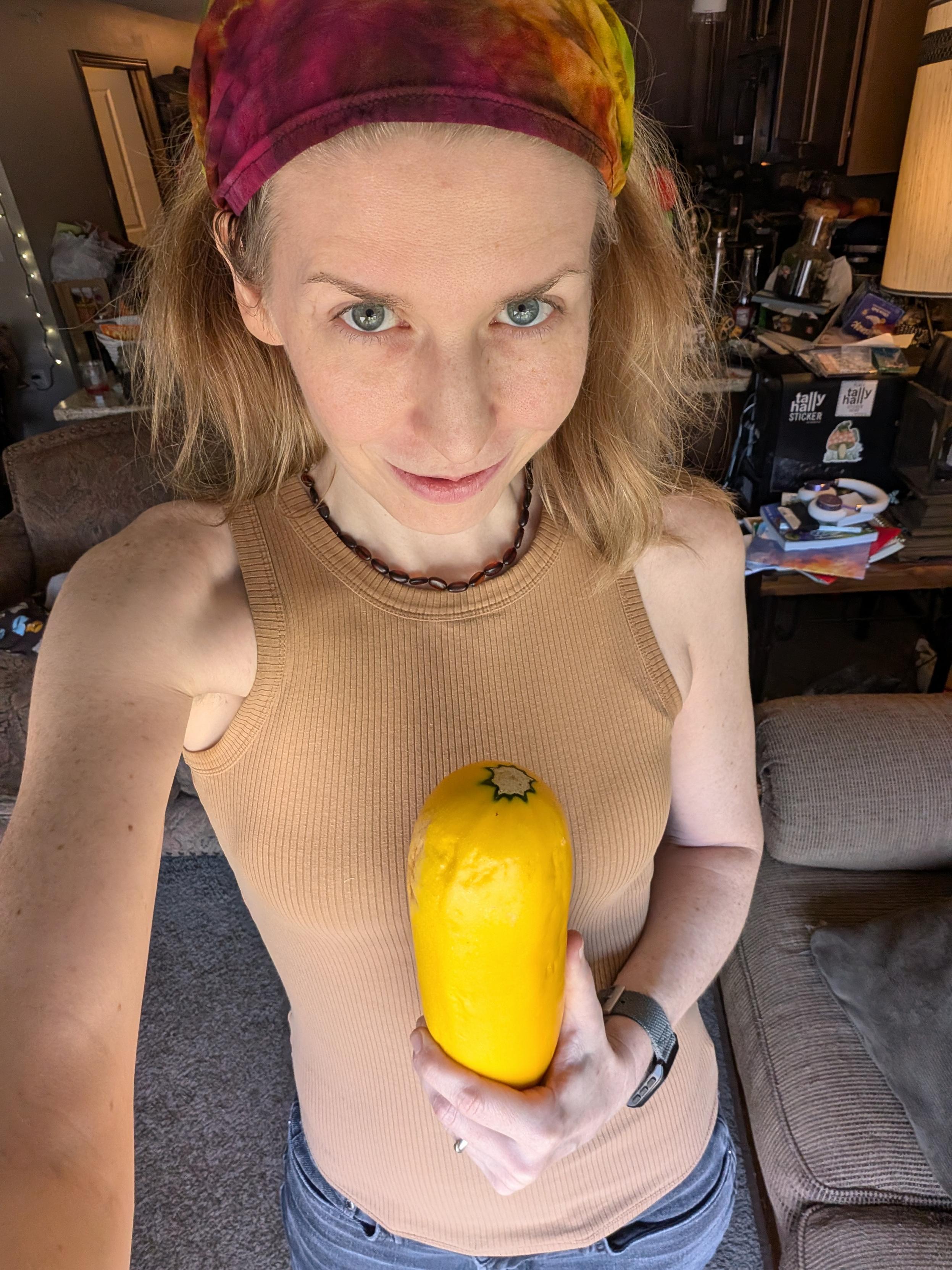 a person with messy long hair held back by a head scarf holds a big yellow....squash...in front of their body, while giving you a Look of some kind