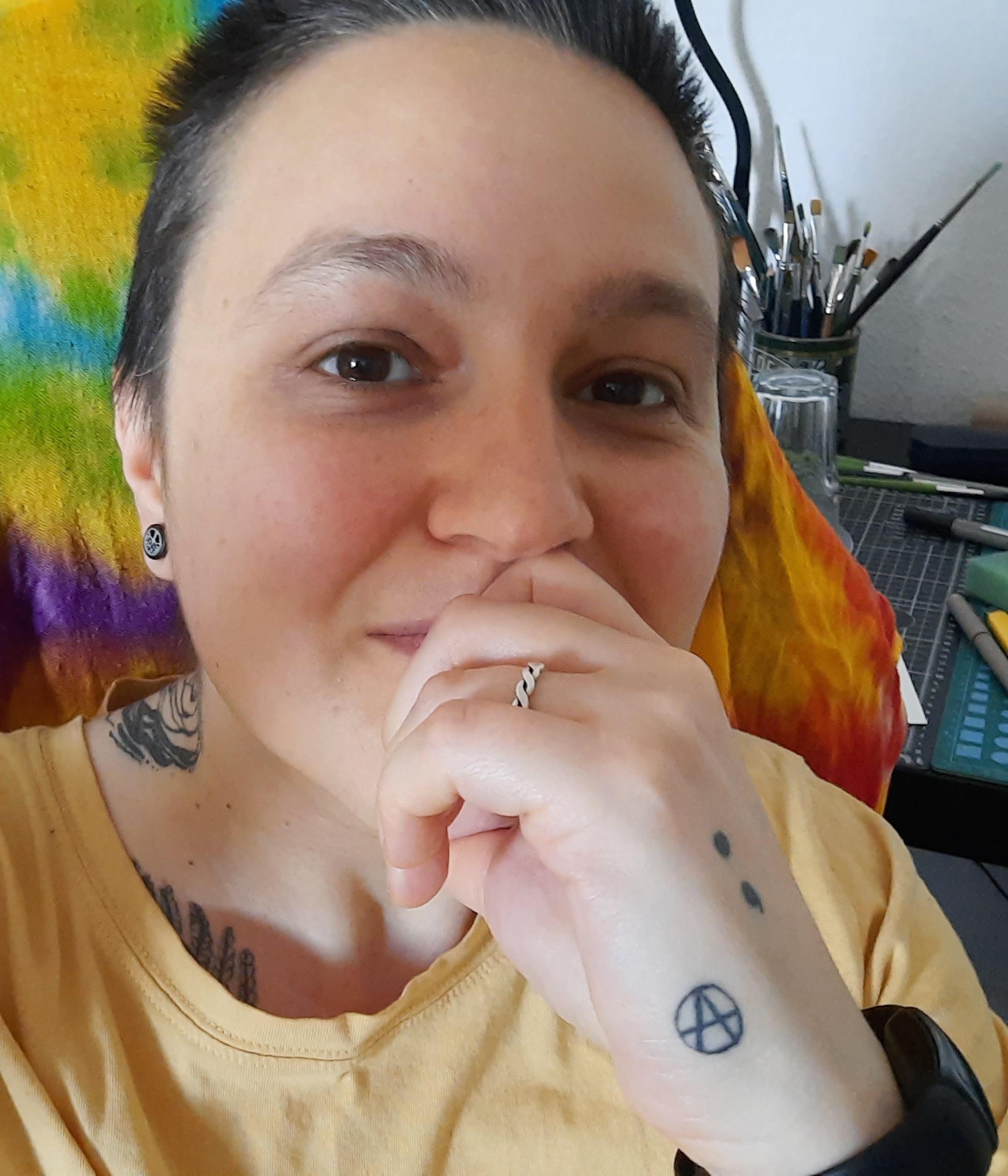 Anxiously posting a pic of myself on social media. Might delete later 😅 Photo of me in an oversized yellow t-shirt that I swear I will eventually make into a Colman's Mustard T-shirt when I have the time to make a stencil. With my hand to my mouth, showing off an anarchist tattoo and a Semicolon tattoo on my hand. Smiling anxiously with a colourful piece of fabric on the chair behind me and evidence that I am an artist in the background (my desk, cutting mat, paintbrushes). I have short brown, scruffy, hair with hints of grey at the front. I look like I just got out of bed to take this photo because after 10 mins looking for a pic, I realised I don't have any without my partner, so yes I did have to get out of bed to take this photo that I might delete later. 🥹 I'm not fond of having my face on the internet these days which is one of the myriad of reasons why I am so bad at selling my work online. I just cannot do all this "selling myself" shite. Reels, talking etc. Bleh. In person I am fantastic and very charismatic but making myself into a product on social media makes me want to vomit. This is problematic because my disability makes selling stuff IRL difficult too. Sigh.