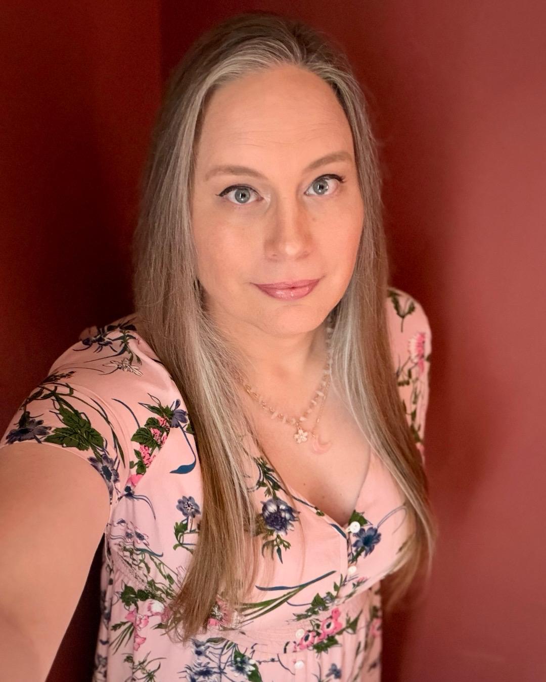 Selfie of a white woman with long, dark-blonde hair and blue eyes wearing a pink floral dress.