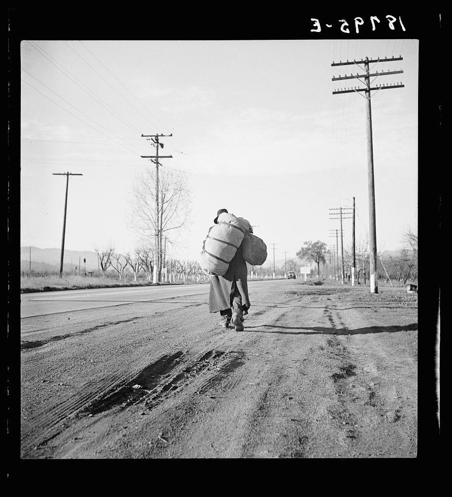 A black and white photograph depicts an individual walking down a rural road, carrying large cylindrical objects on their back. The person is dressed in light-colored clothing with dark boots, suggesting possible work attire suitable for labor-intensive tasks. They are seen from behind as they traverse the dirt path marked by tire tracks. Telephone poles line either side of the road and barren trees punctuate the background under a clear sky. There's an air of solitude around this figure, highlighting their journey or task ahead in such an environment. The image is captioned with "3-29P81," which could be indicative of its date or cataloging information. It carries historical significance as it references Carleton Parker and the Industrial Workers of the World (IWW), indicating that Dorothea Lange, known for her work during the Great Depression documenting laborers' lives, captured this moment. This photograph is part of a larger narrative on California's rural landscape and its connection to industrial workers. The mention of Napa Valley places it in relation to the region's agricultural history.