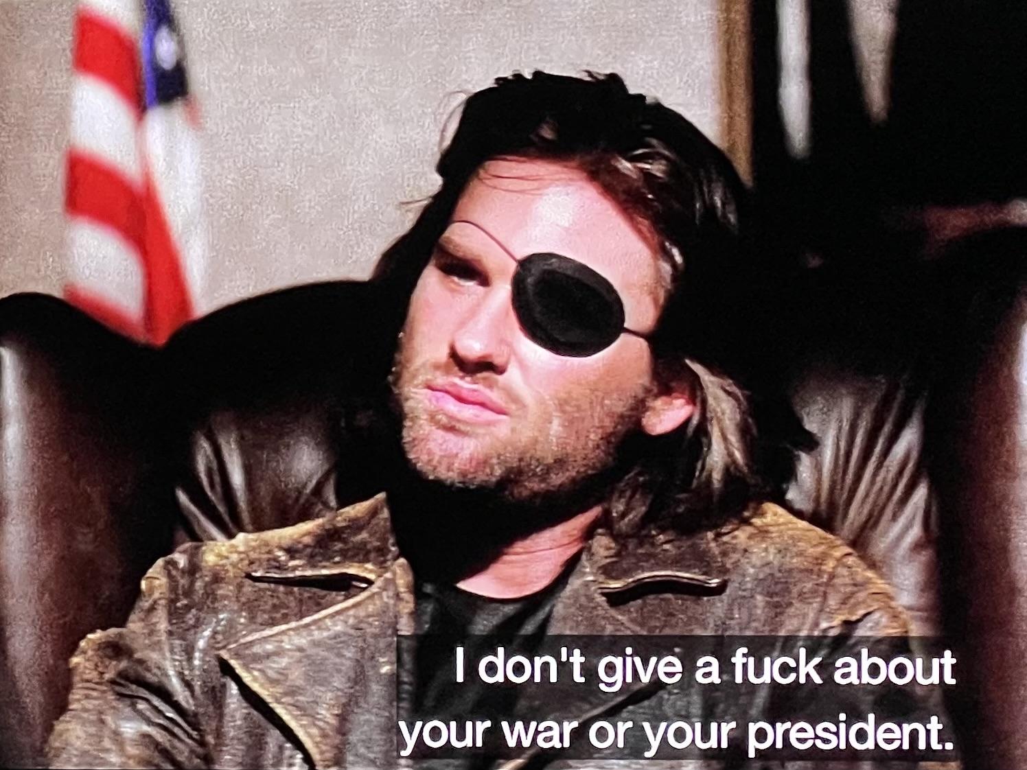 Kurt Russell as Snake Plisskin in Escape From New York (1981). He’s seated in a leather wingback chair wearing a very weathered leather jacket with his signature eye patch and fantastic hair saying ‘I don't give a fuck about your war or your president.’