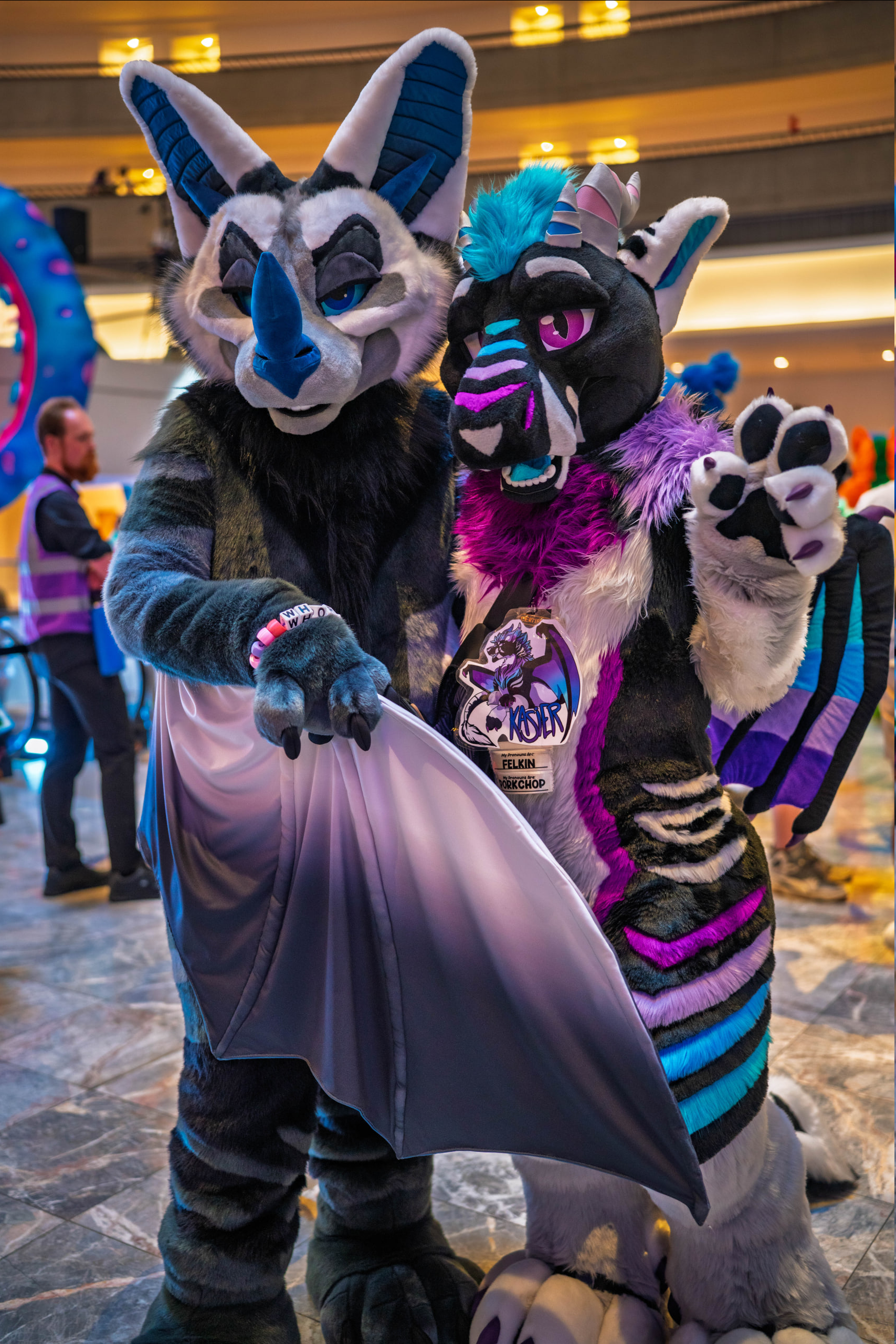 A photo of a bat fursuiter wrapping a felkin fursuiter in his wings while the felk waves