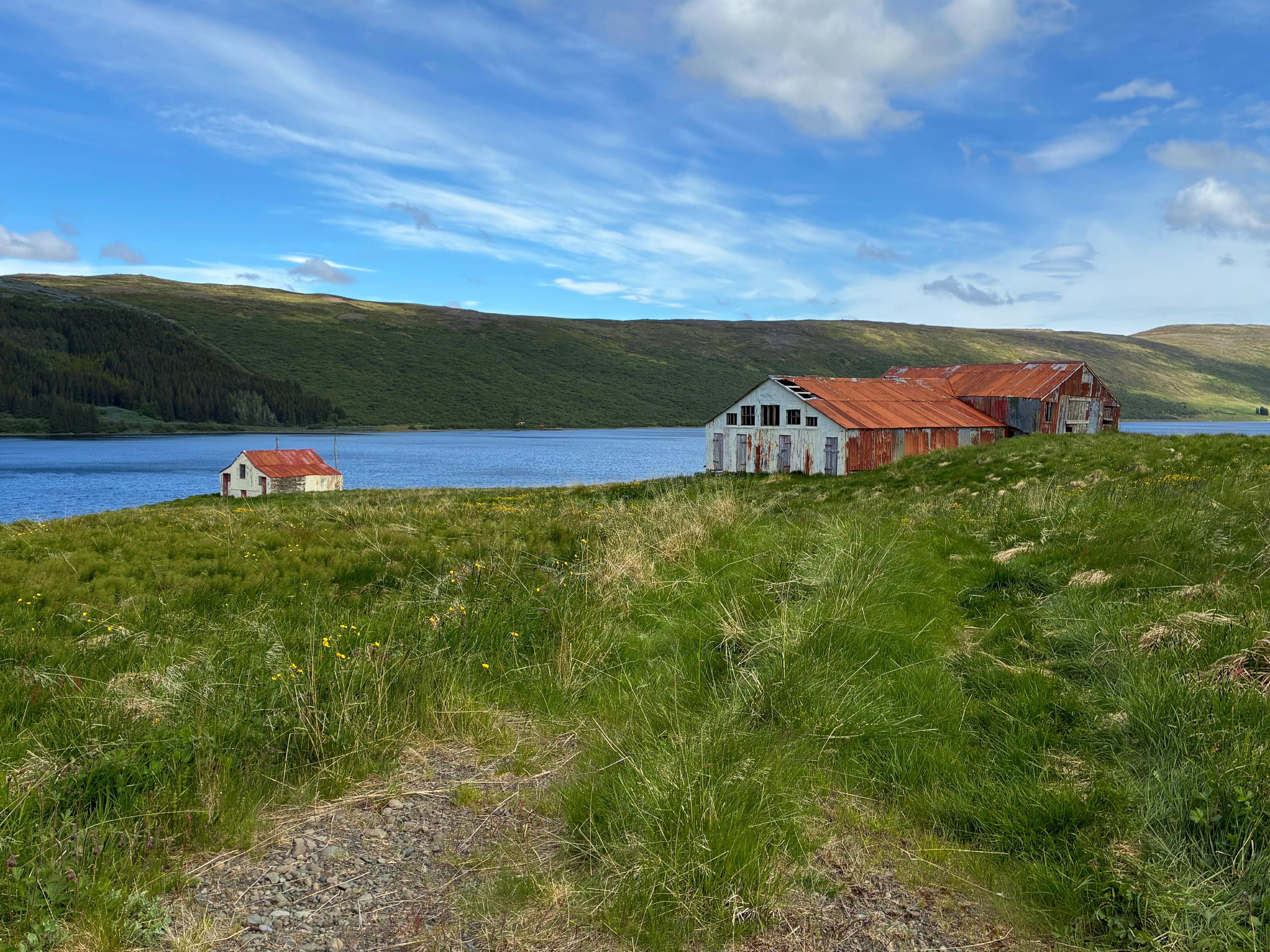 A small abandoned house next to a rusty barn with a lake and a mountain in the background.
