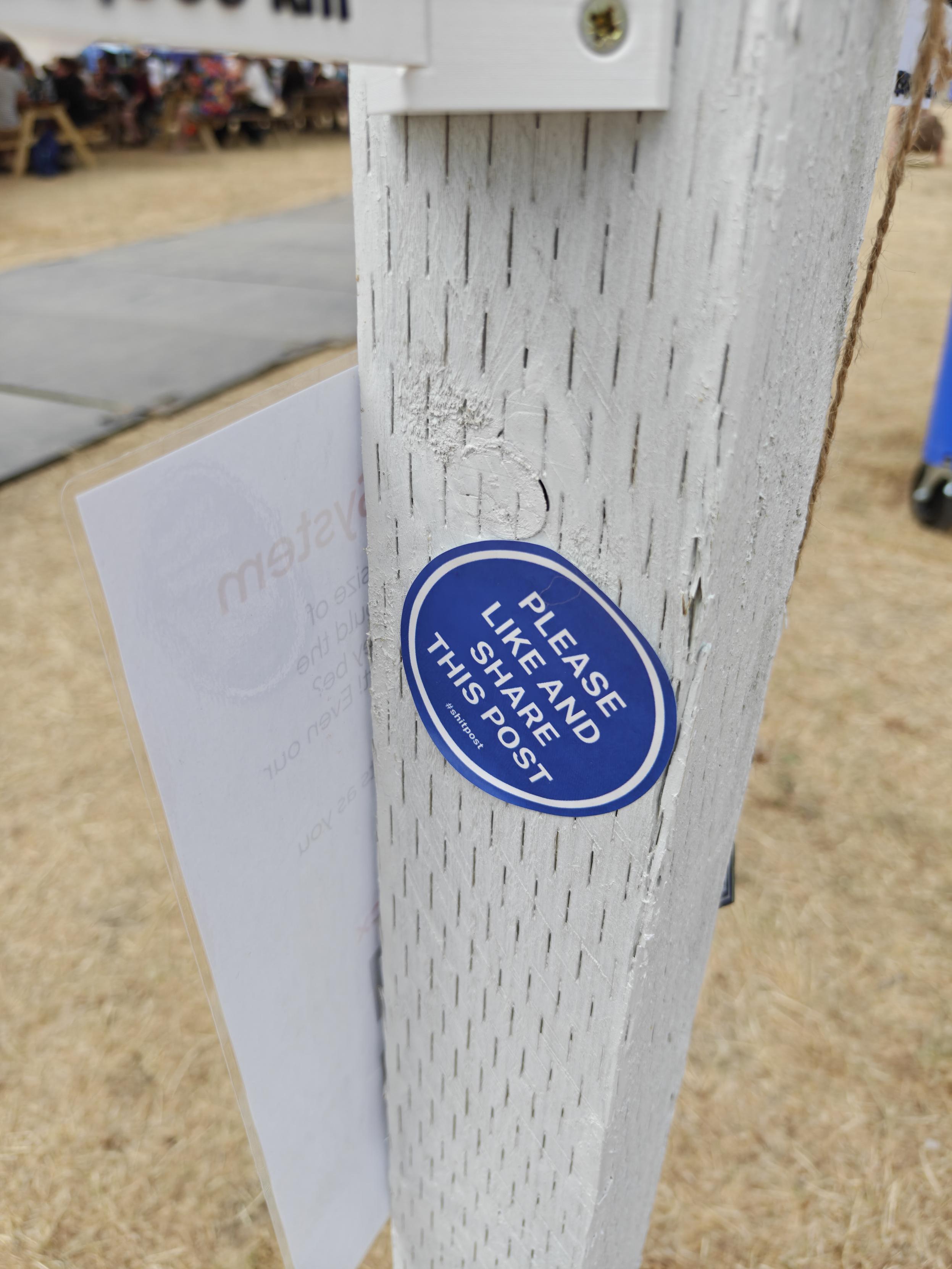 A white post. On it is a blue sticker saying "PLEASE LIKE AND SHARE THIS POST"