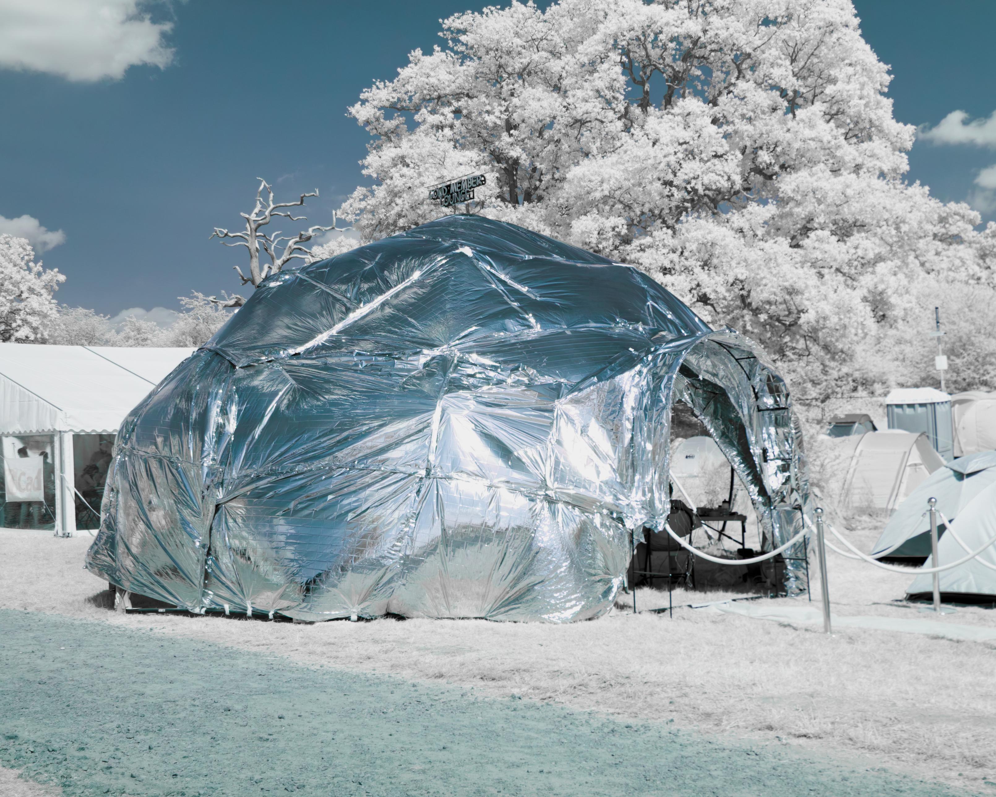 a dome tent in infrared, under a blue sky and a bright white tree. it is surrounded by other tents and trees. all is pale and glowy