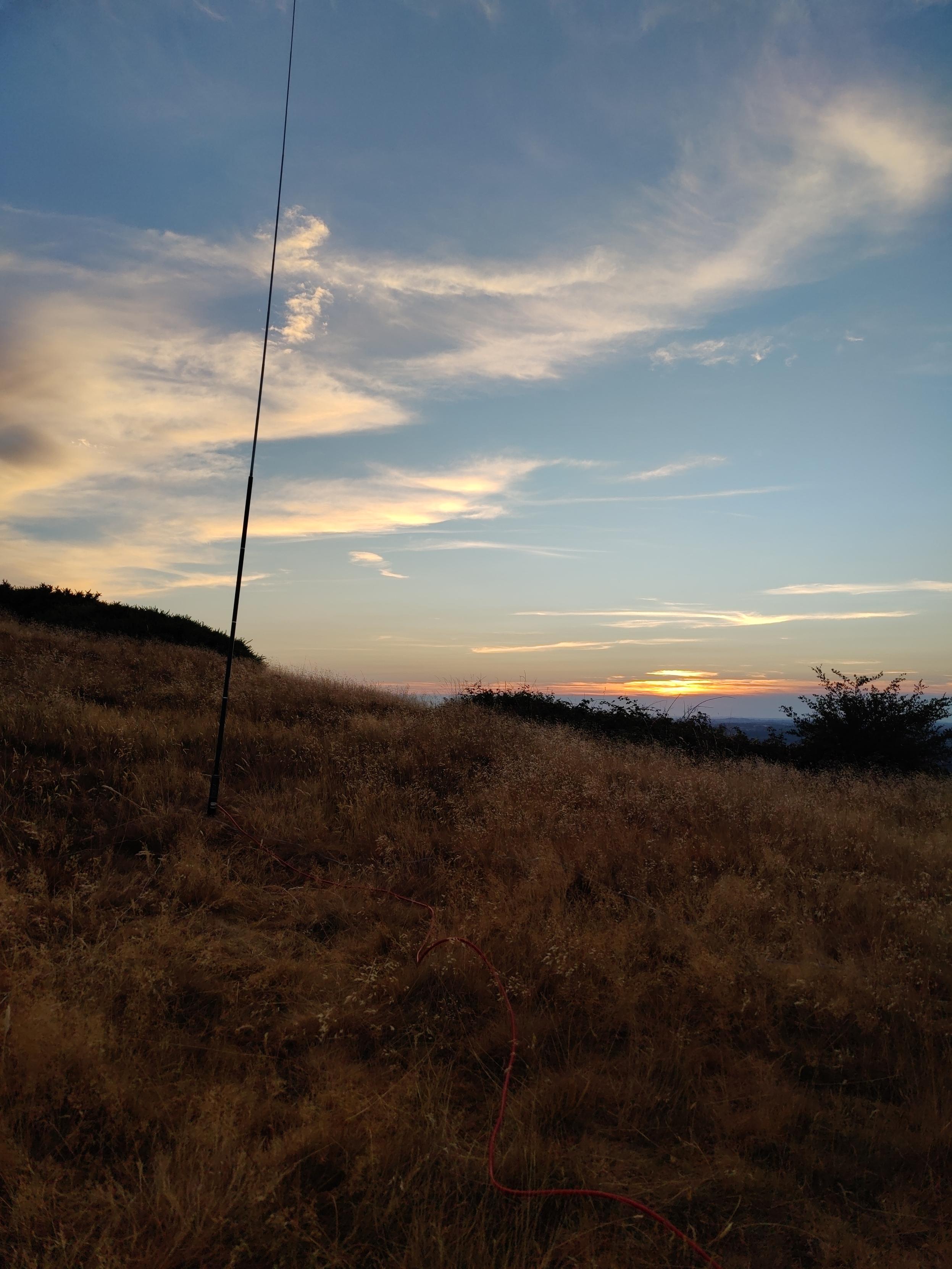 Looking towards sunset, the sun is low and obscured by a layer of cloud but the sky is mostly clear and blue with some strokes of white and orange clouds. In the foreground a dry grasses and other flora with some bushes along the sloping ridge of the hill we're on. A vertical antenna is shoved into the ground and reaches out of the top of the frame, you can make out the red coax snaking back to our POV