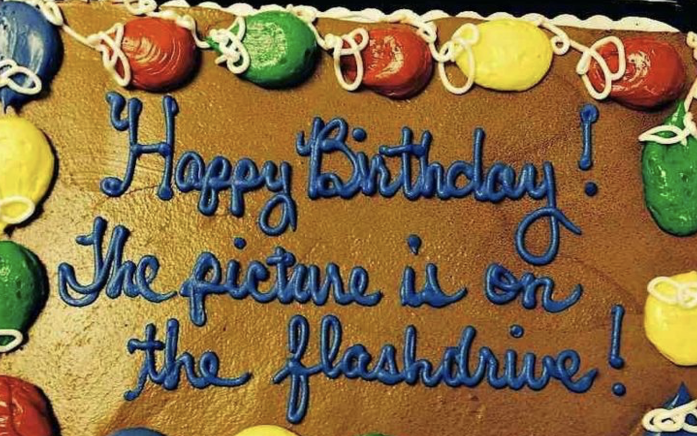 A cake with icing balloon decorations and the text "Happy Birthday! The picture is on the flashdrive!" written across the top in blue icing