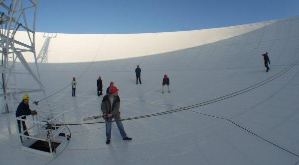 Several people standing in the large 76m dish of the Lovell Telescope. The person nearest the camera has a cricket bat stood near the centre of the bowl. Four people are fielding. One person nearer the edge of the dish is bowling.