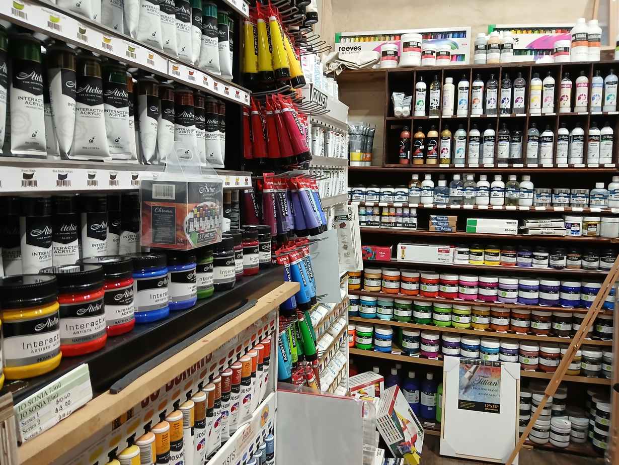 Corner shelves in the art shop, with the backwall at the back of the photo and the wall shelves down the left side. The wall shelves include hanging tubes of paint, and shelves of paint jars. The back walls have more jars, and bottles of paint in little cubby holes. It's packed full of colours, all of the colours.