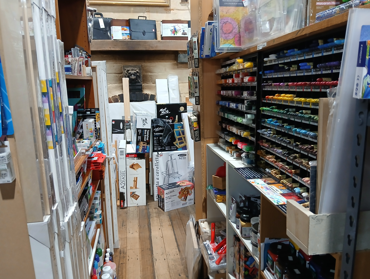 Looking down one of the narrow aisles toward the back wall. The back wall is hidden behind boxed easels in all sizes, a shelf above them holds small folio cases. The right wall of the aisle is oil pastels, with block printing ink on the bottom shelves. The left side is lined with stretched canvases and boxes of pastels and pencils.