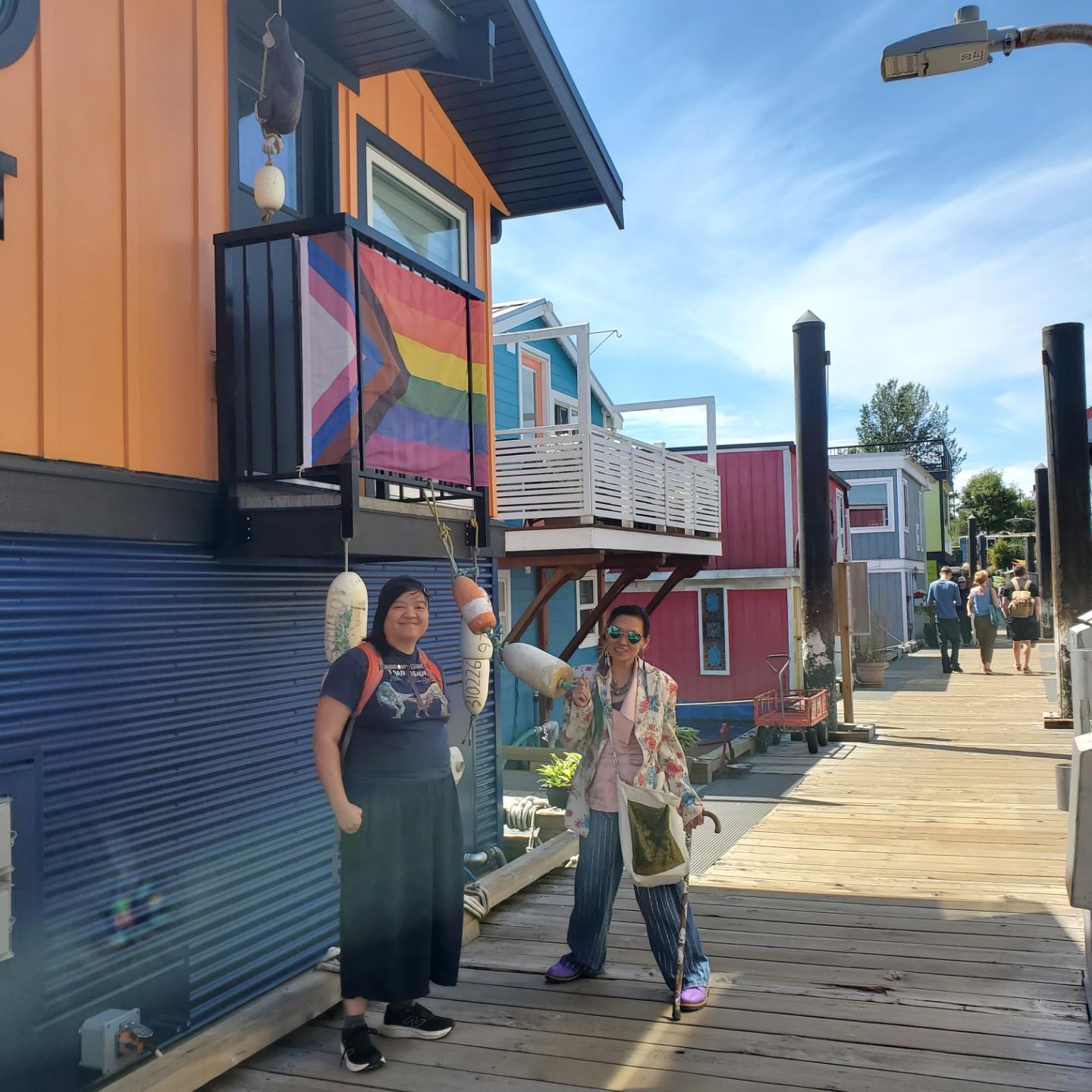 Me & May stand on a wooden boardwalk next to a row of colorful floating homes. A Pride & Trans flag hangs on the orange floating home on the left.