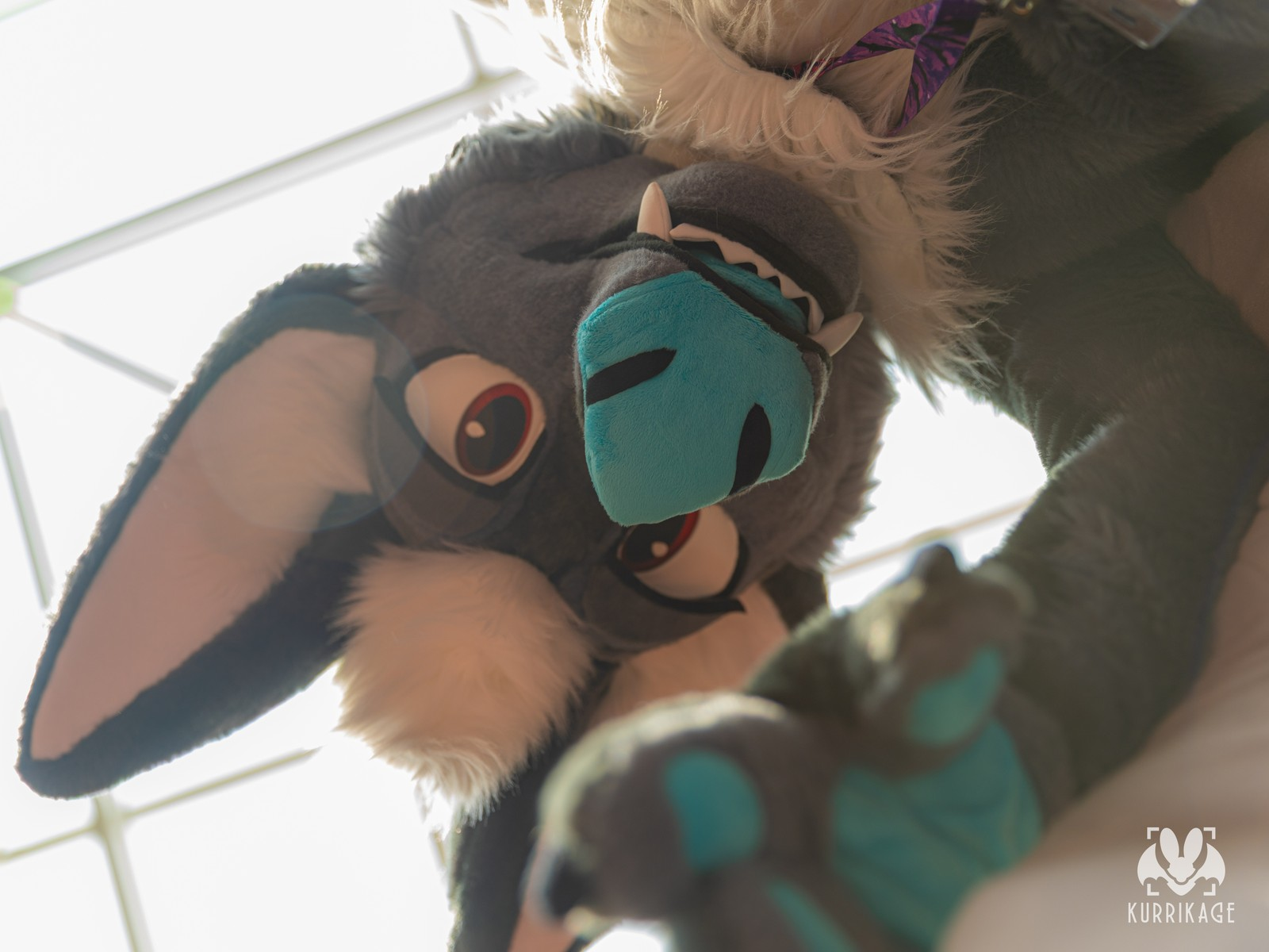 A low angle view of a bat fursuiter reaching down towards the viewer below against the blinding light of a skylight.