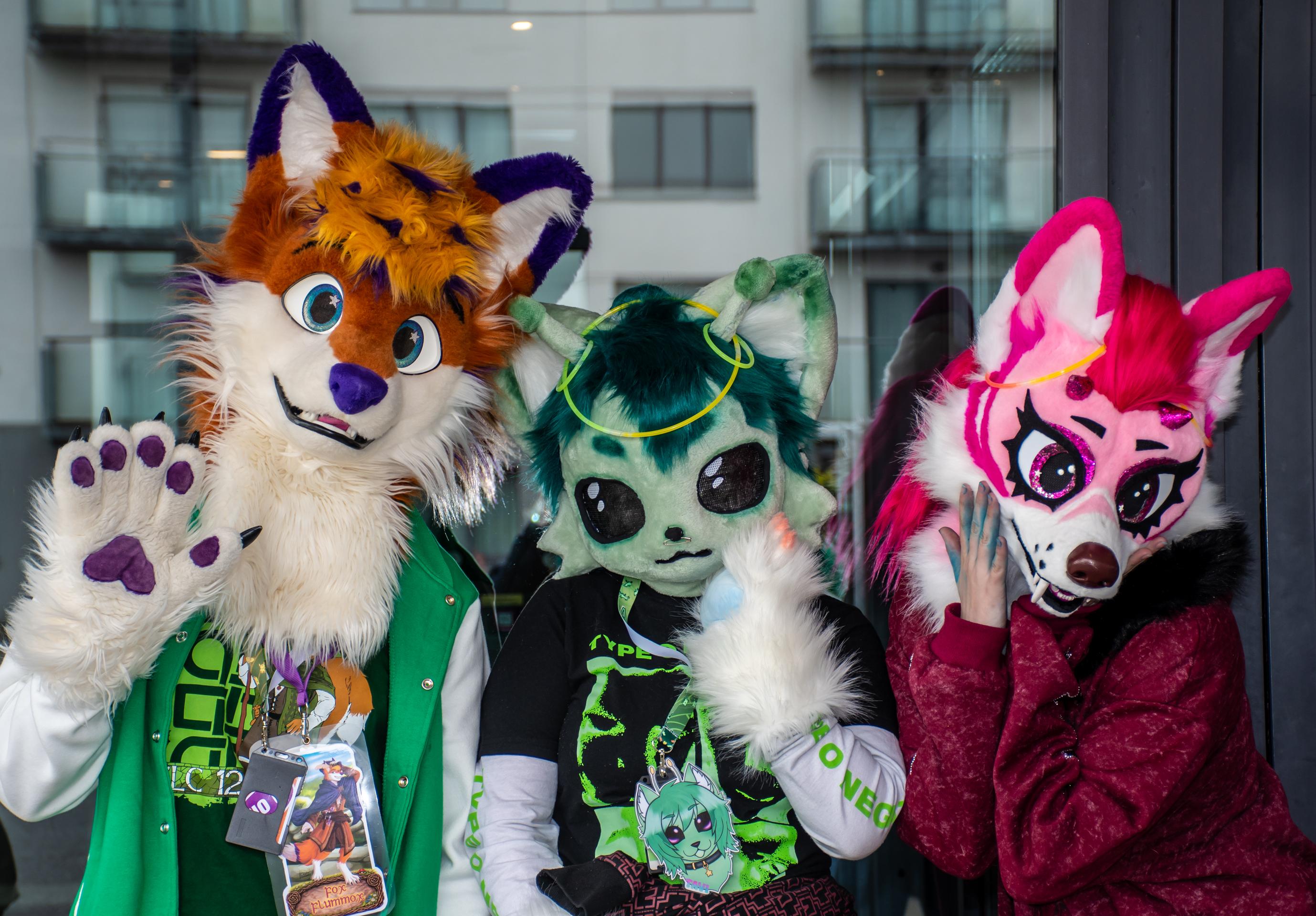 A portrait with 3 partial fursuiters. On the left Fox Flummox wearing a green con jacket and themes badges, waving into the camera. In the middle the "kemono alien" Tavi with big black insect-like eyes and little antennae. On the right side pink sabertooth Pixie with lots of glitter.