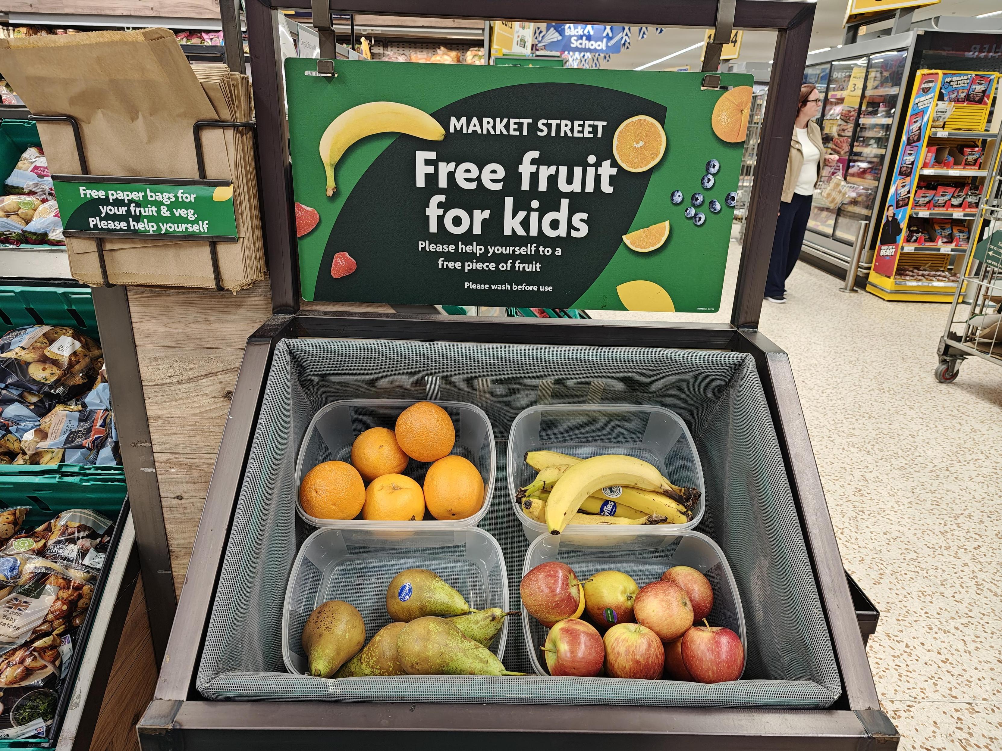 A supermarket container with punnets of oranges, banana, pears and apples on display, with a sign saying "Free fruit for kids - please help yourself to a free piece of fruit" above them.