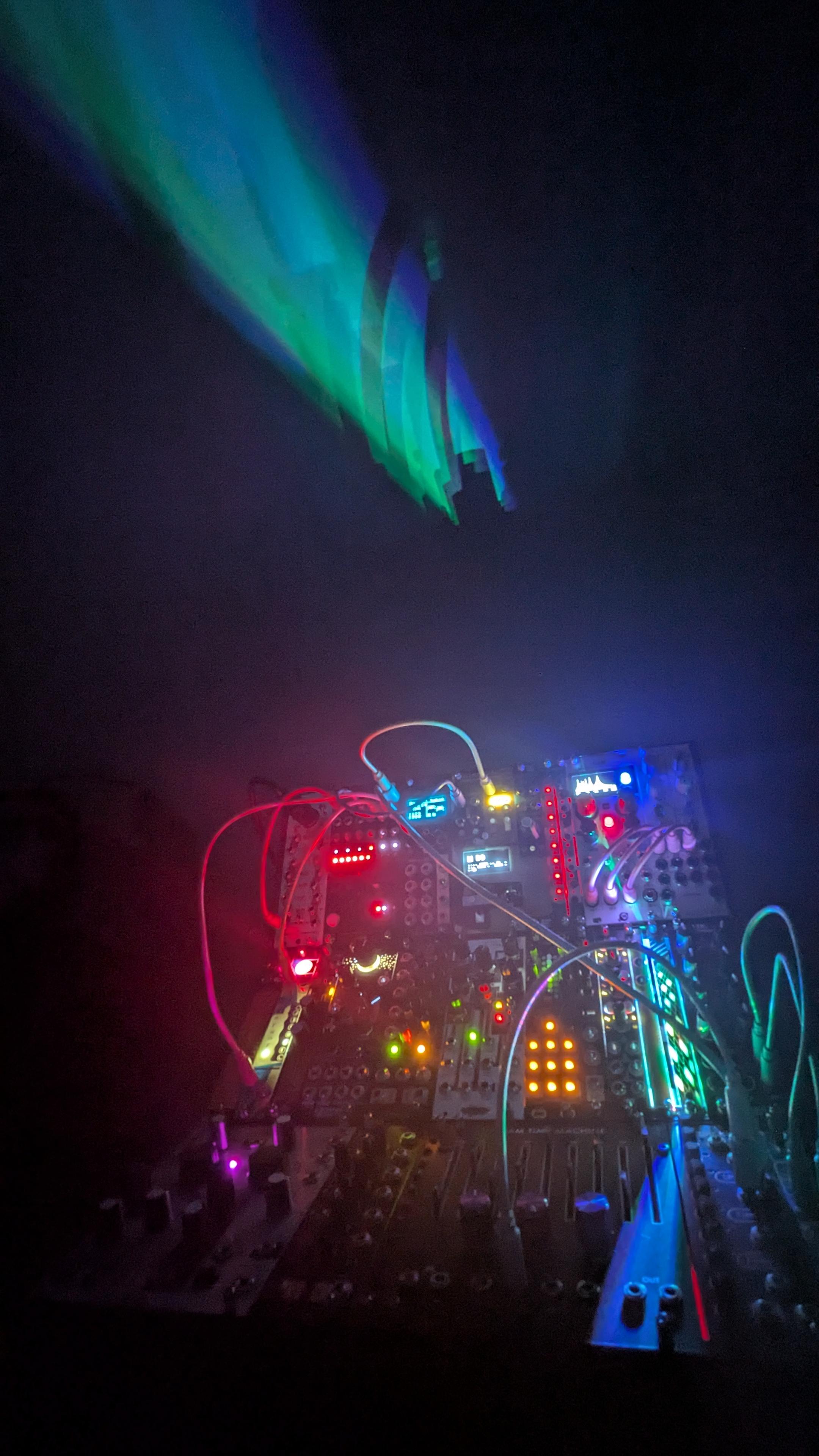 A large modular synthesizer in a dark room. the many LEDs light up the room with many colors. A strip of color hits the ceiling from a gap between the modules
