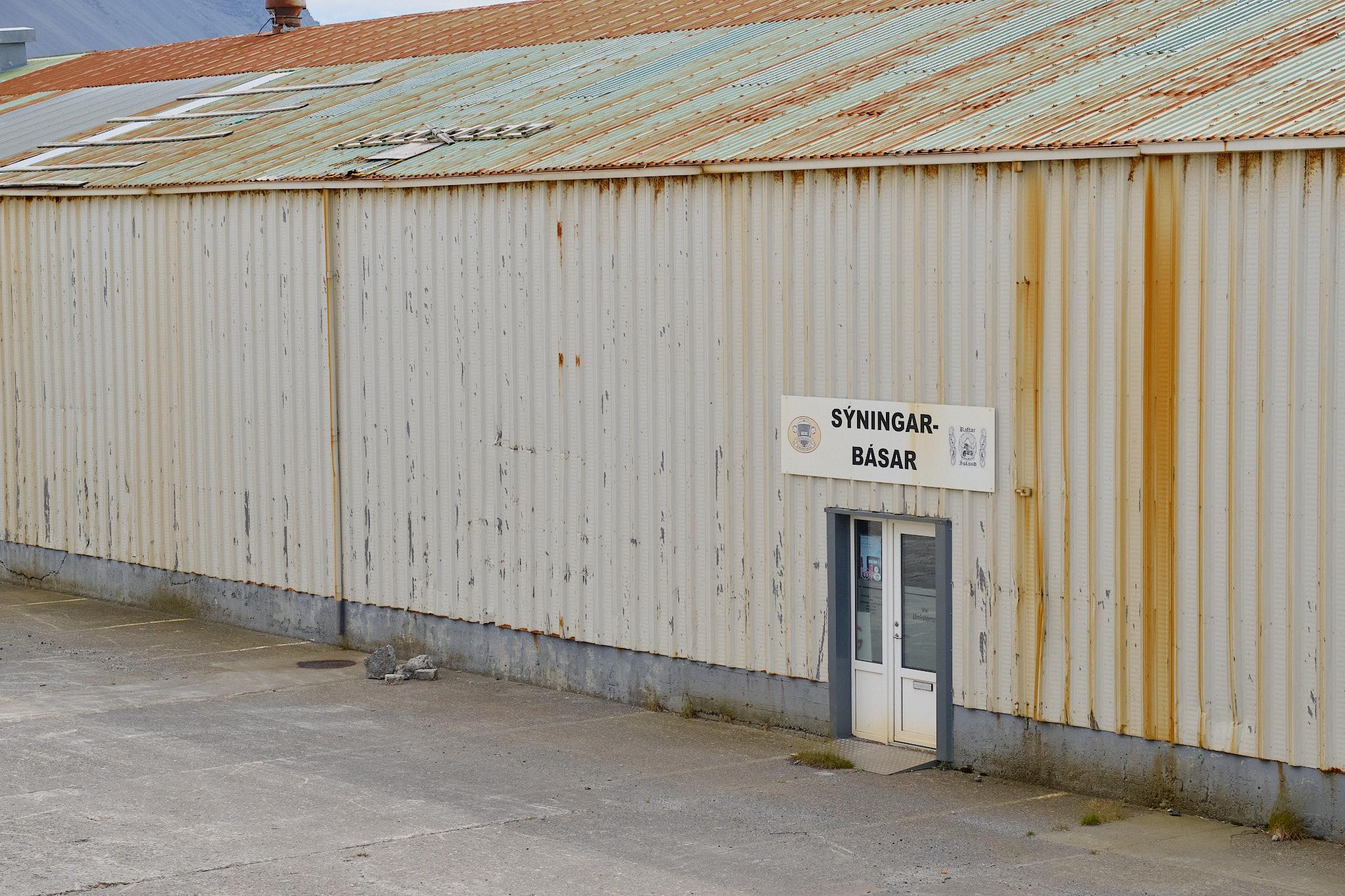 An old sign over an old door on a rusty corrugated-iron wall. The sign says "sýningarbásar", or "show booths", and "Fornbílafélag Borgarfjarðar" or "the antique car association of Borgarfjörður". The building is now unused.