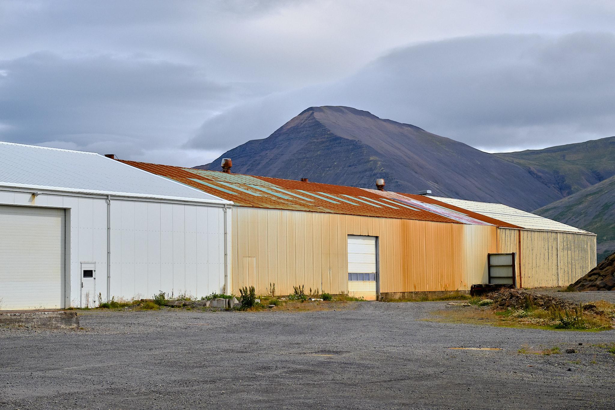The other side of that large warehouse, showing the rusty corrugated iron. A mountain towers over it in the background.