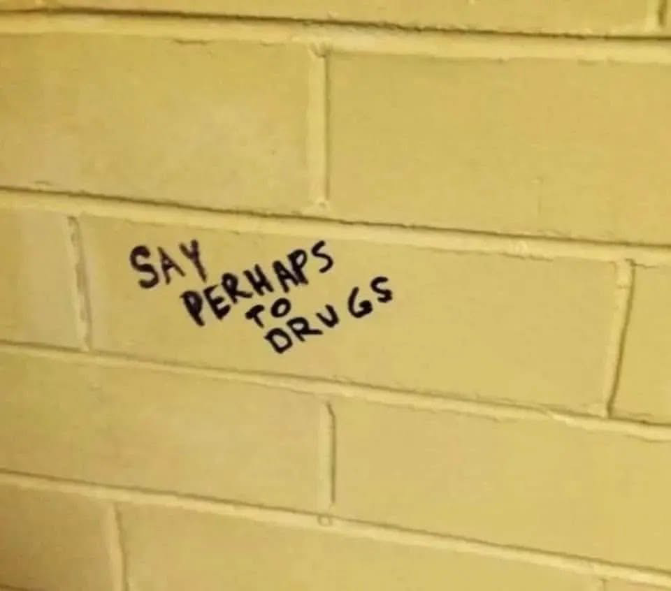 A section of yellow-painted cinder block wall, the kind you would find in a public bathroom. Written on it in black capital letters, at a slant, it says "SAY PERHAPS TO DRUGS".
